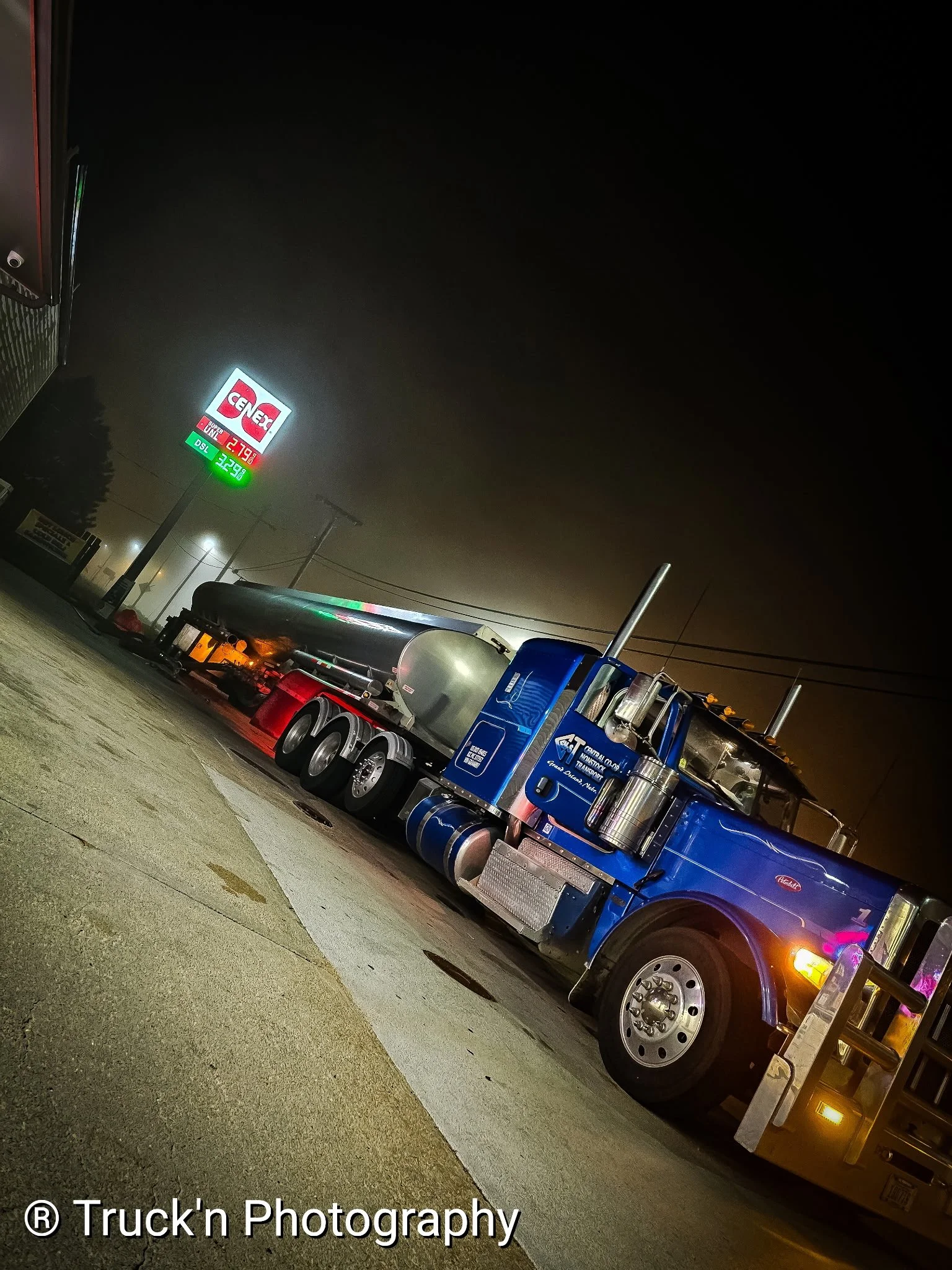 Nighttime photo of a blue semi-truck parked at a gas station with a large sign displaying fuel prices, including regular gasoline, diesel, and LPG.