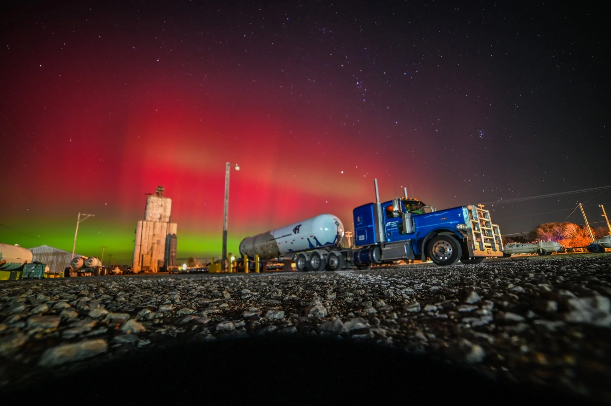 Nighttime scene with a blue semi-truck and trailer parked on rocky ground, under a sky illuminated by colorful northern lights, stars visible in the background, and some industrial buildings and utility poles nearby.