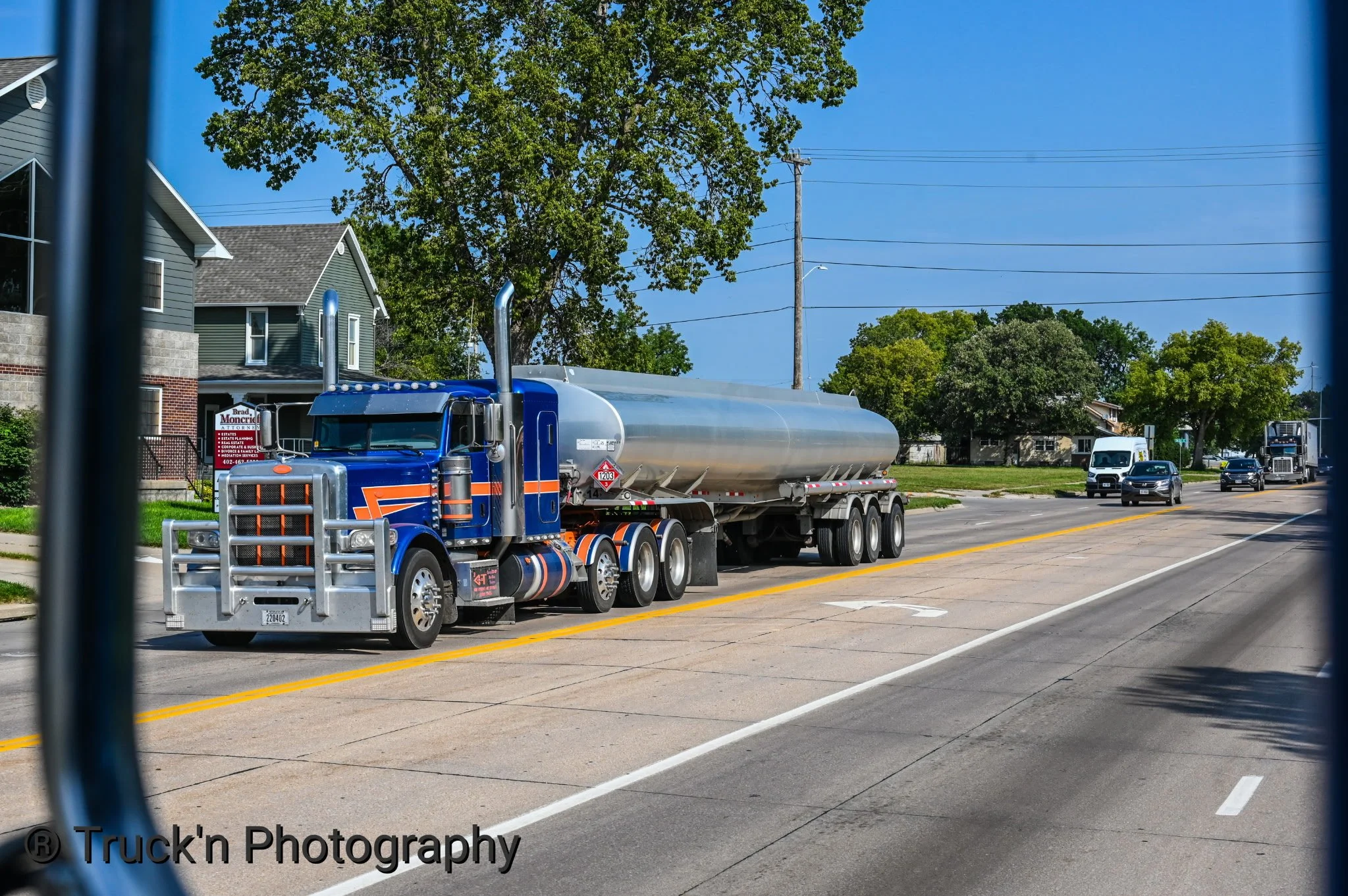 A blue semi-truck hauling a large metal tank trailer on a city street with residential buildings and trees in the background.