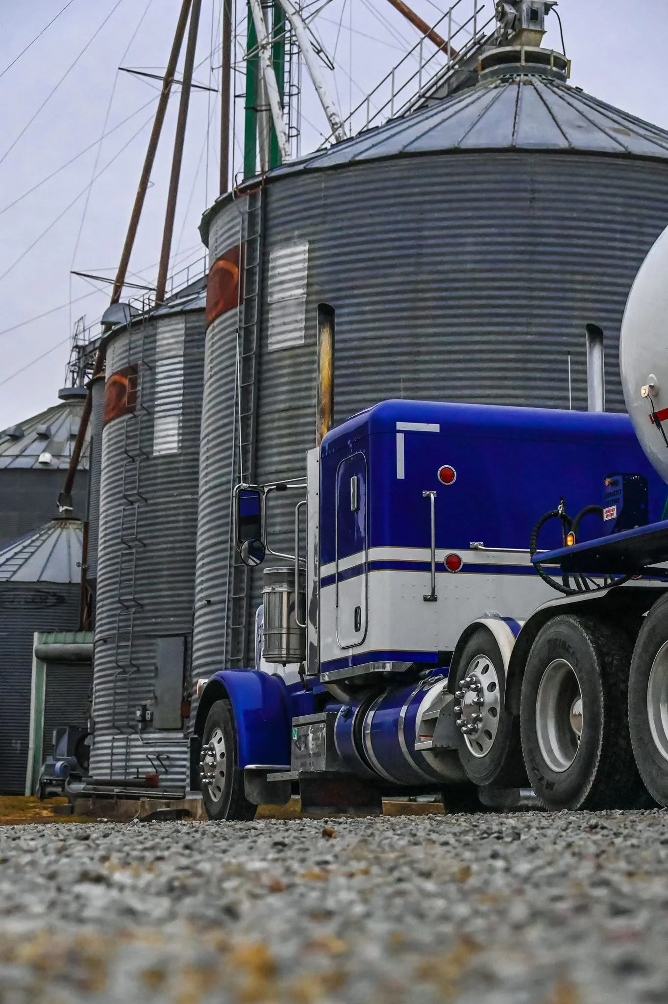 A blue and white semi-truck parked in front of a large metal grain silo on a gravel surface.