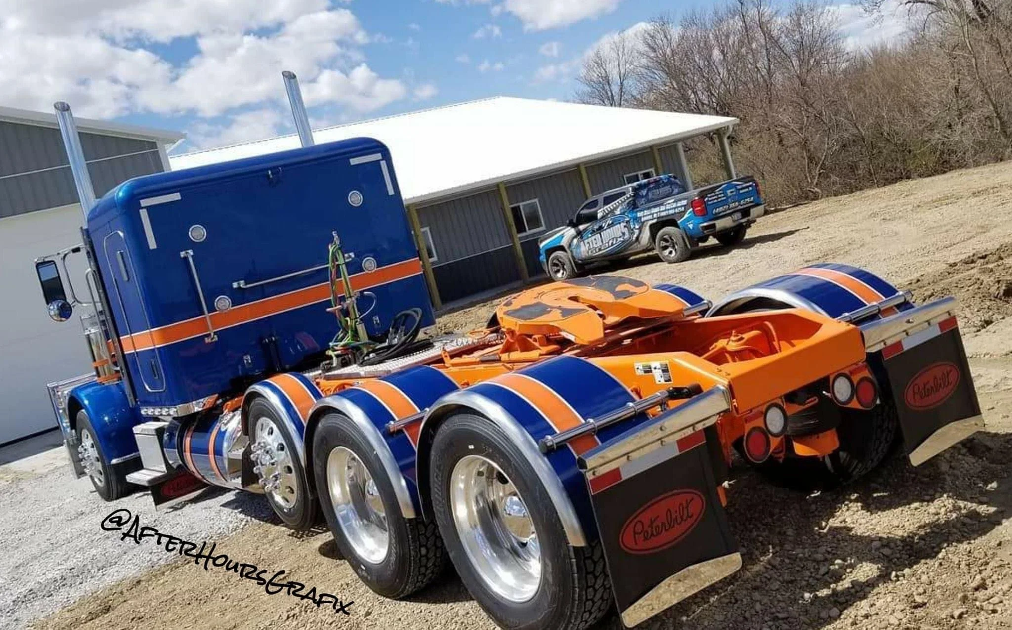 A blue and orange semi-truck tractor cab without a trailer parked on a dirt lot with a warehouse building and a blue patrol vehicle in the background.