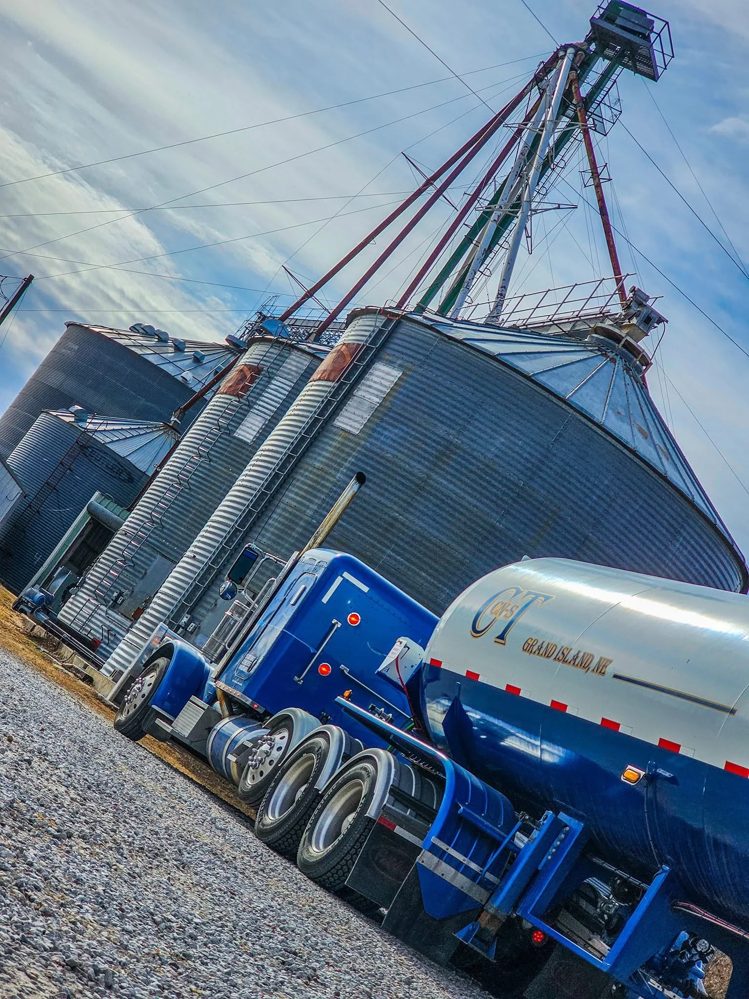 A large industrial grain silo with a blue semi-truck parked in front of it on gravel ground under a cloudy sky.