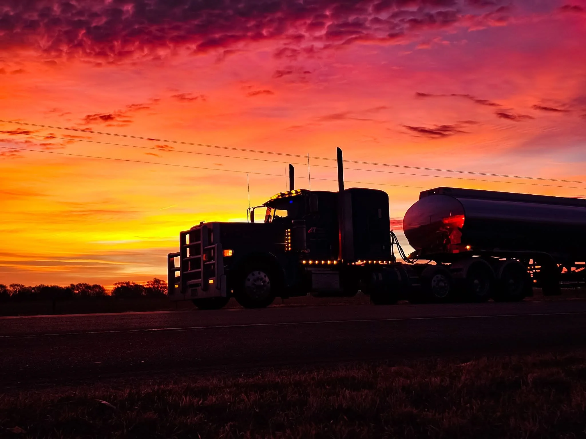 Silhouette of a semi-truck with a tank trailer against a vibrant sunset sky with pink, orange, and purple clouds.
