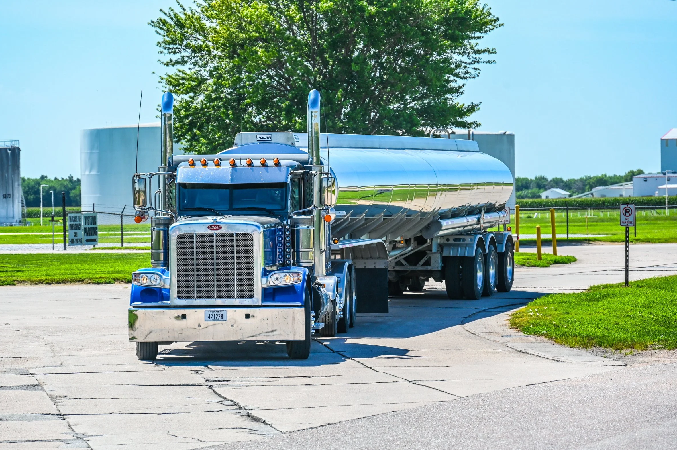 A large blue tanker semi-truck driving on a paved road with a green field and trees in the background.