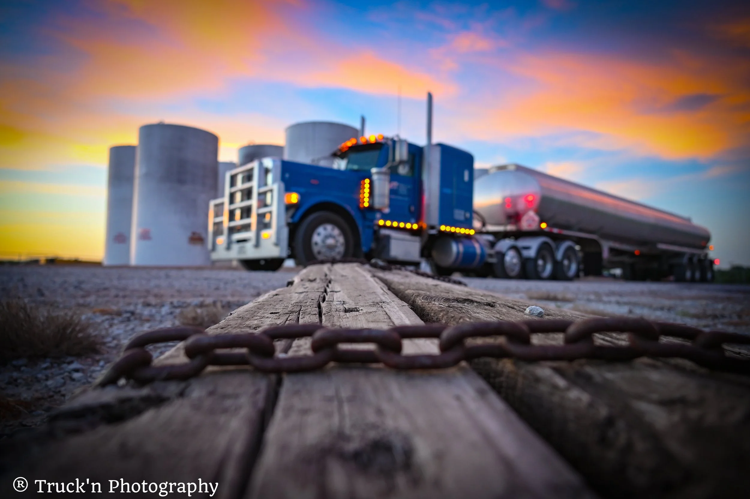 A shot of a blue semi-truck on a gravel lot at sunset, viewed from a low angle with a rusty chain in the foreground and storage tanks in the background.