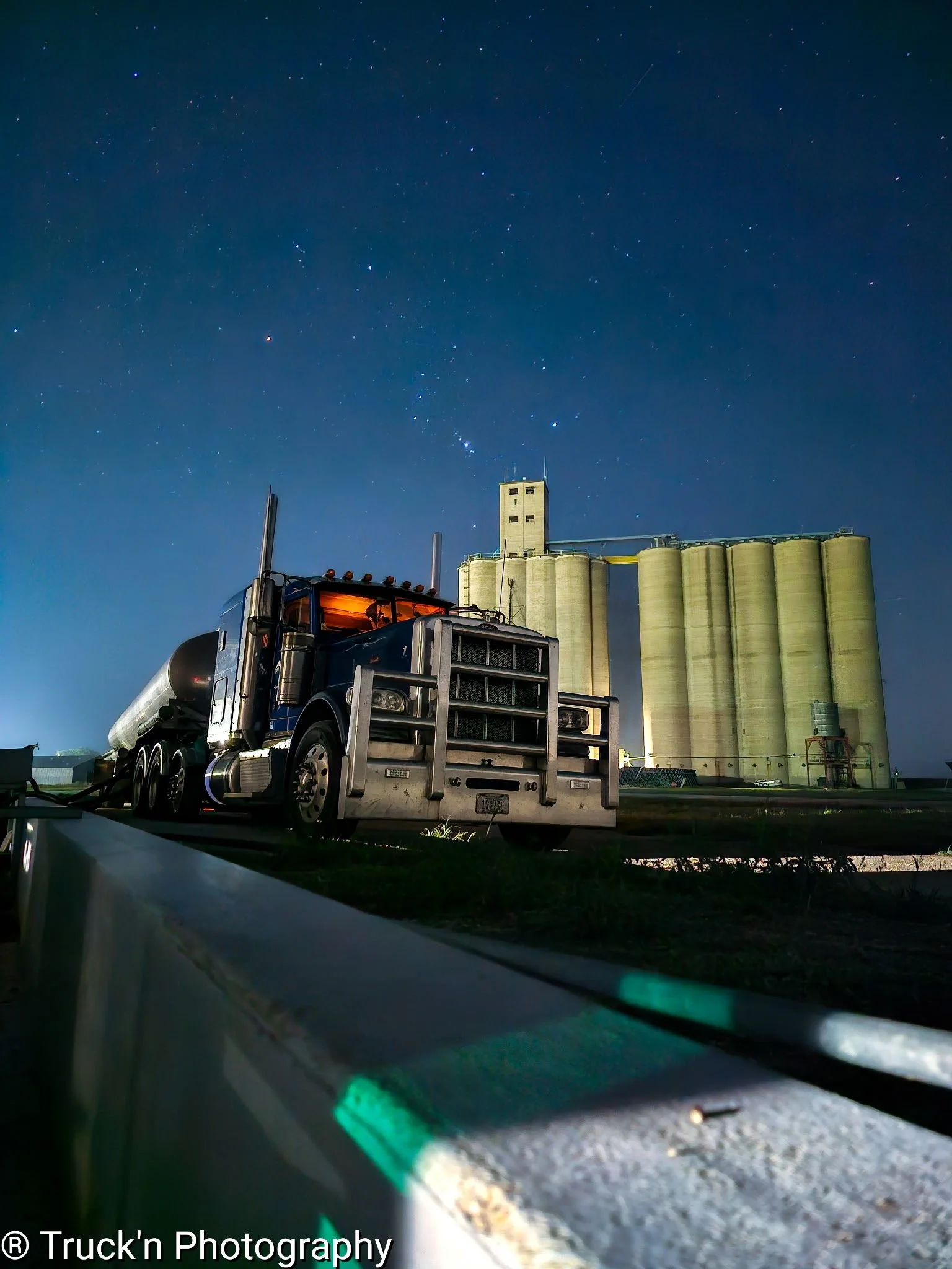 Nighttime scene with a large semi-truck parked in front of grain silos under a starry sky.