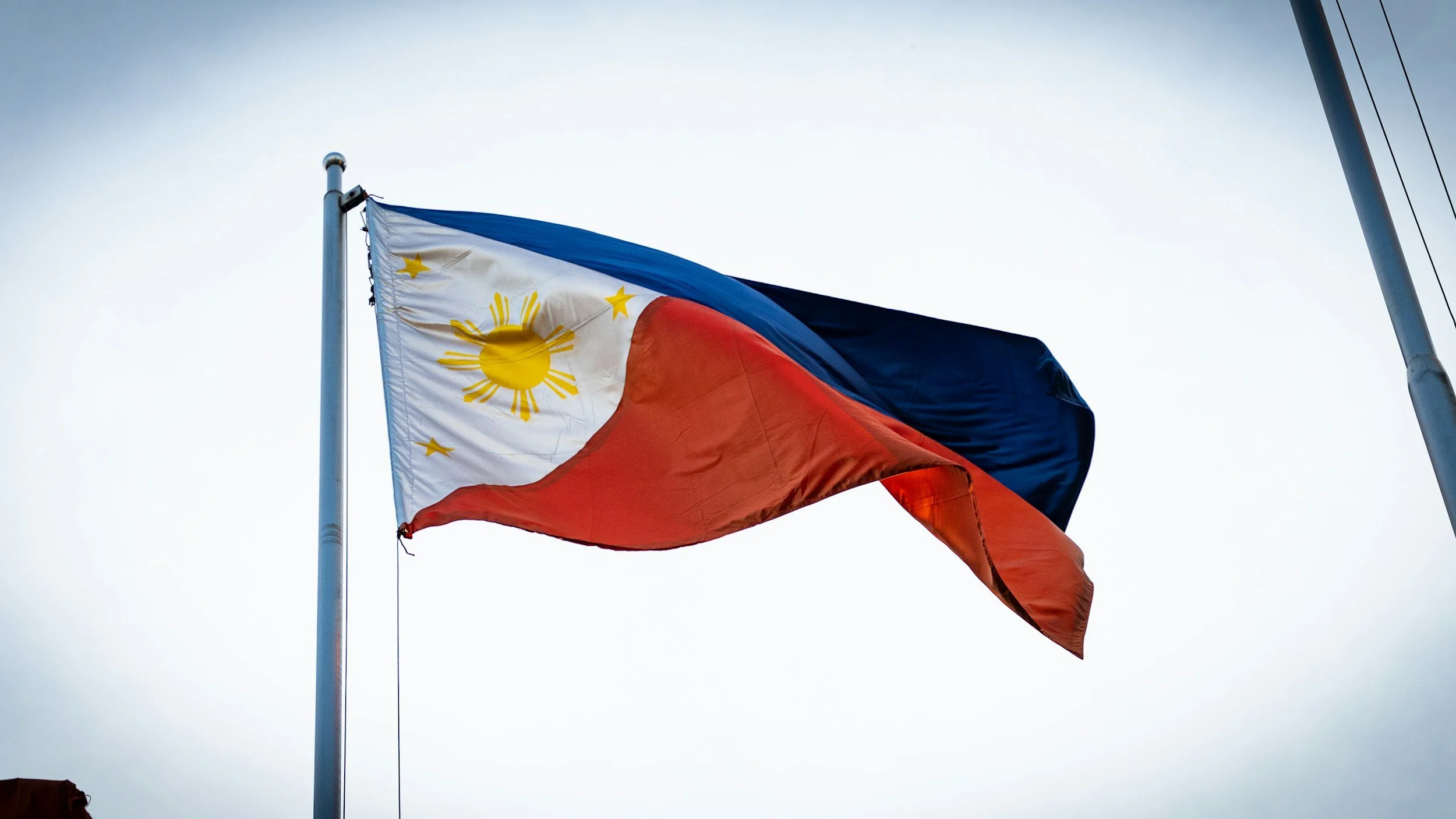 The Philippine flag flying on a flagpole against a cloudy sky.