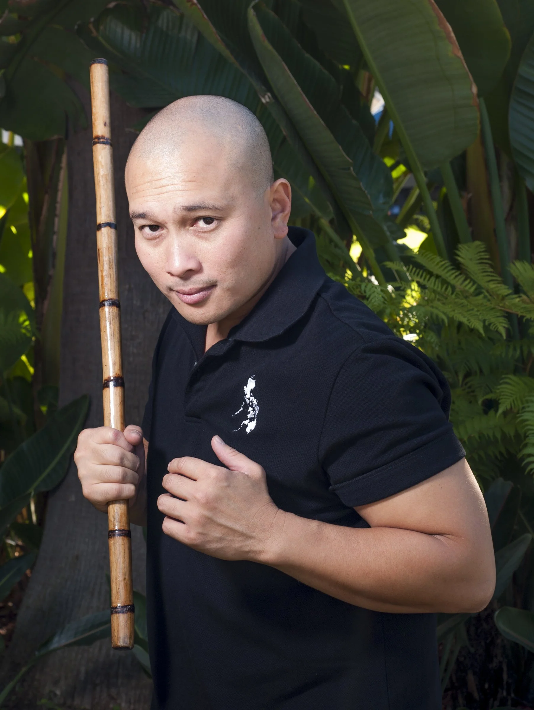 A man with a shaved head holding a bamboo stick, standing outdoors among lush green plants.