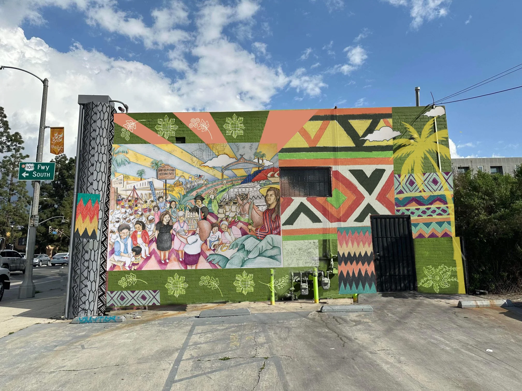 Colorful mural painted on a brick building wall depicting a lively scene with people, artwork, patterns, and nature symbols such as flowers and leaves.