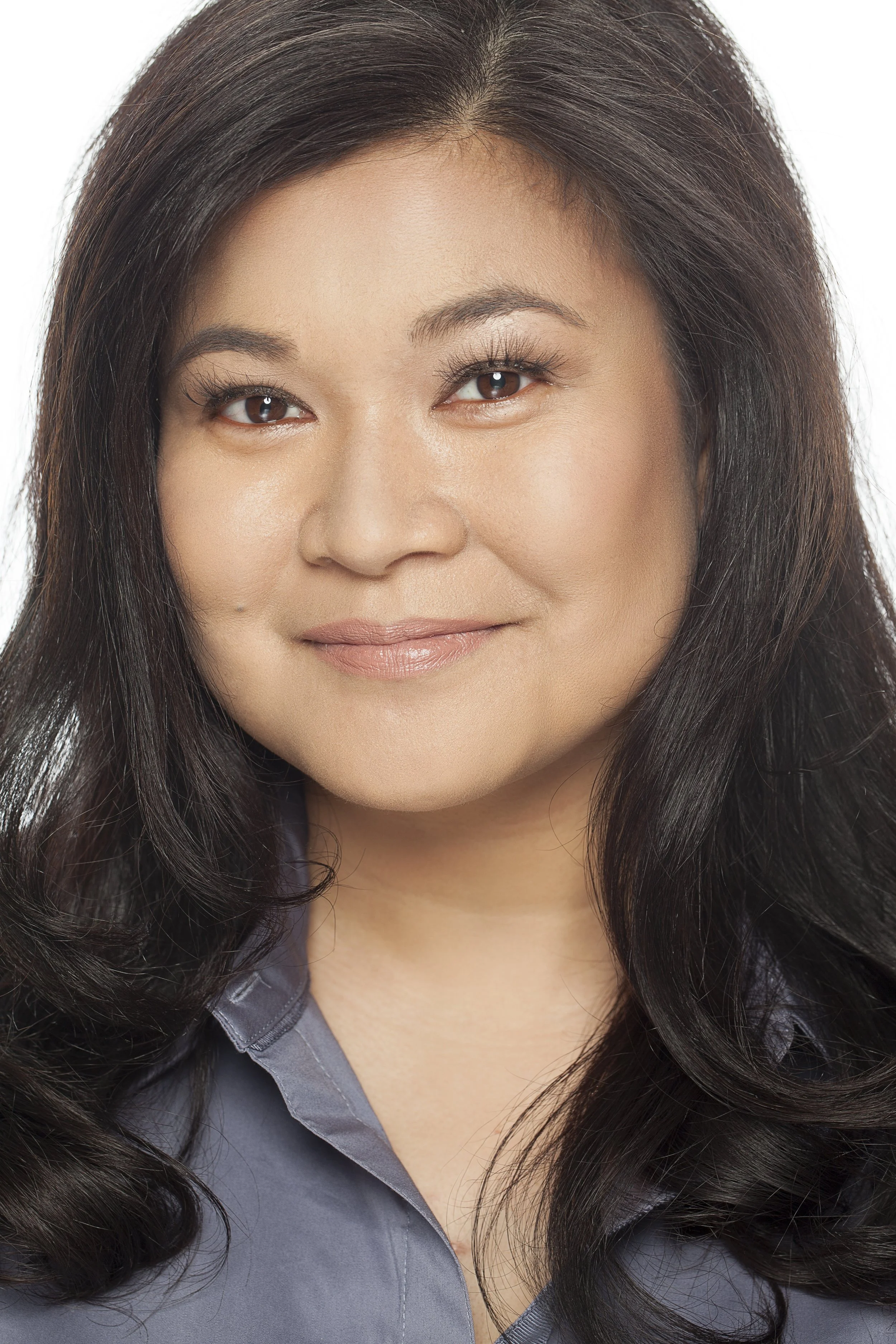 Close-up of a woman with long, dark, wavy hair wearing a gray collared shirt, smiling softly.