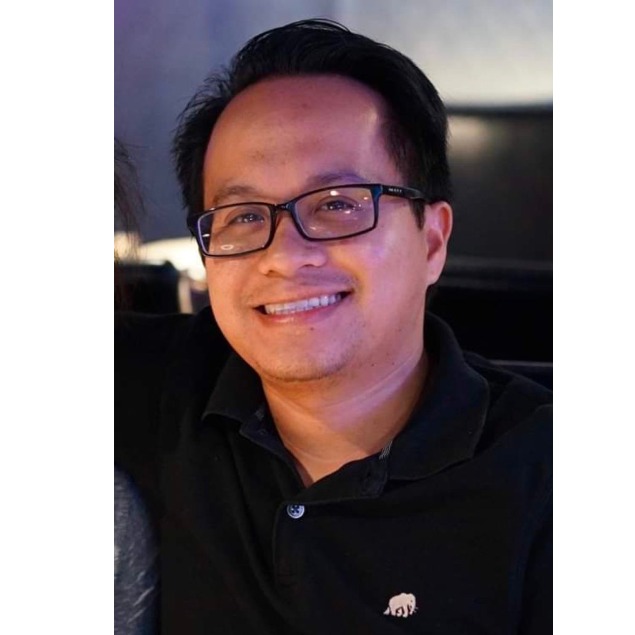 Close-up of a smiling man wearing glasses and a black jacket with a small elephant pin on the lapel.