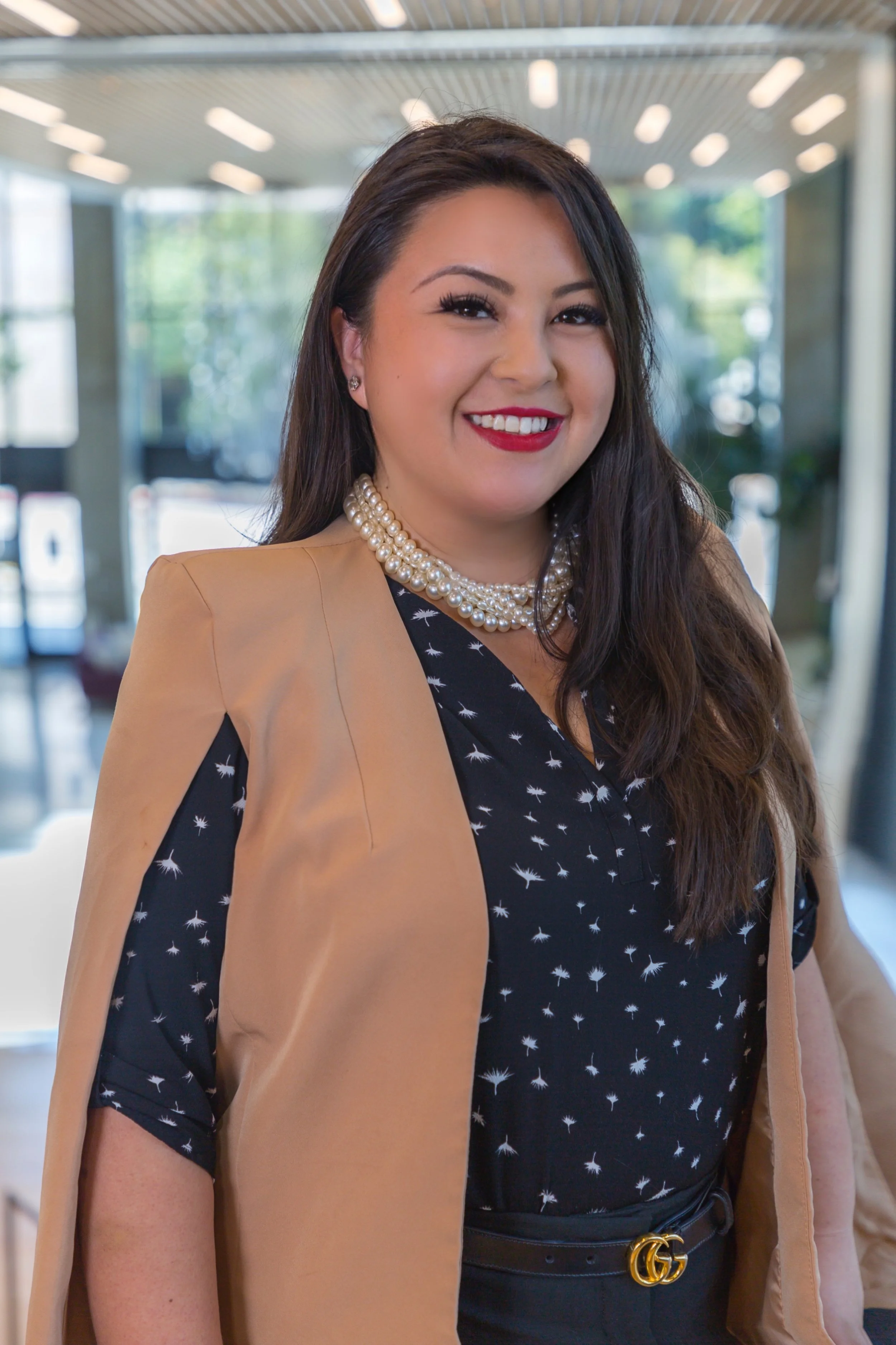 Close-up of a smiling woman wearing a pearl necklace, a beige blazer, and a black blouse with a white pattern. She has long dark hair and makeup with red lipstick, standing indoors with blurred background lighting.