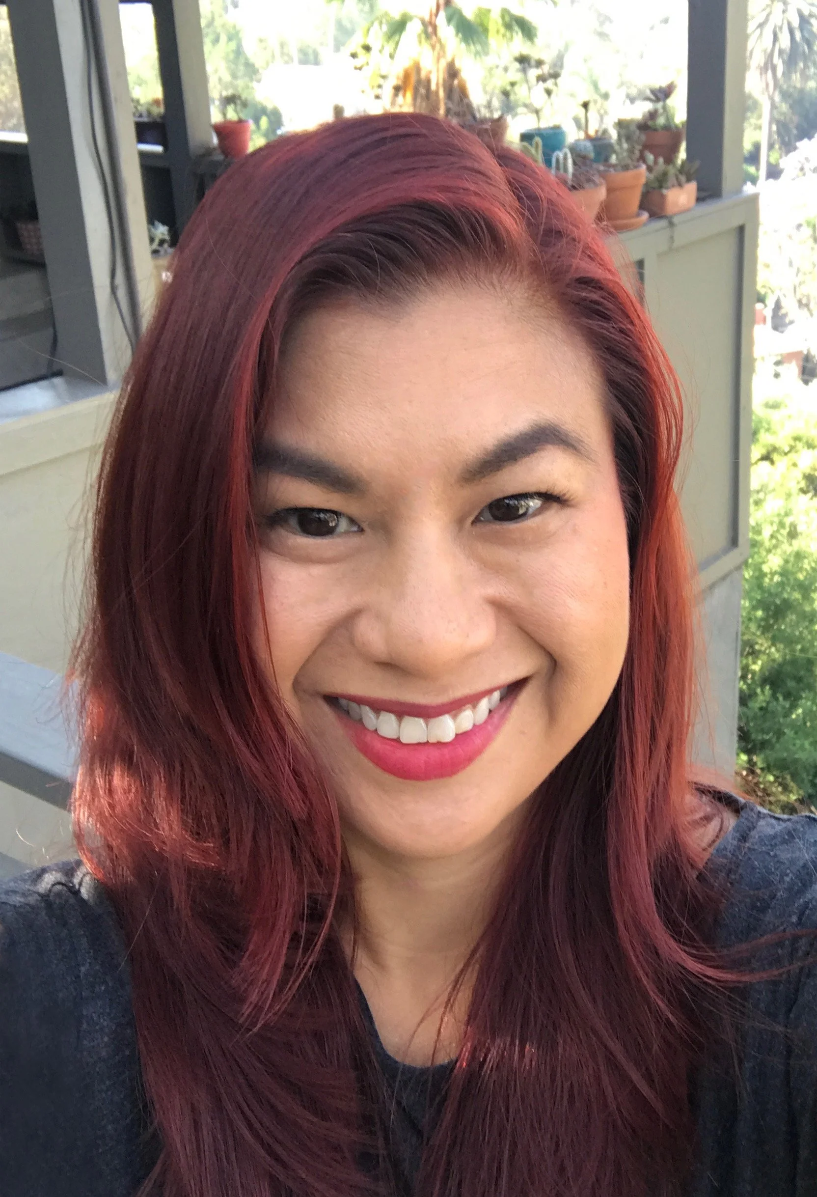 A smiling woman with red-brown hair and makeup, taking a selfie outdoors on a balcony with potted plants and trees in the background.
