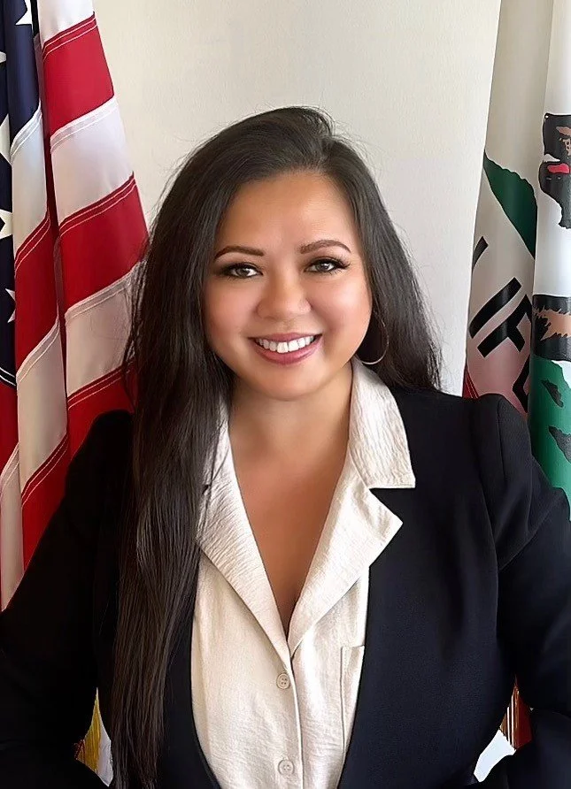 A woman with long dark hair smiling, wearing a beige shirt and black blazer, seated in front of several flags including the United States flag and the California state flag.