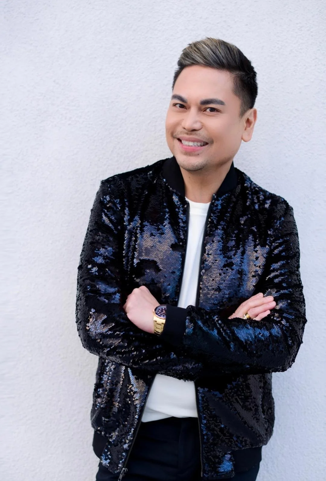 A smiling man with short dark hair, wearing a shiny black and blue sequin jacket, white shirt, and gold watch, standing with arms crossed in front of a plain white wall.