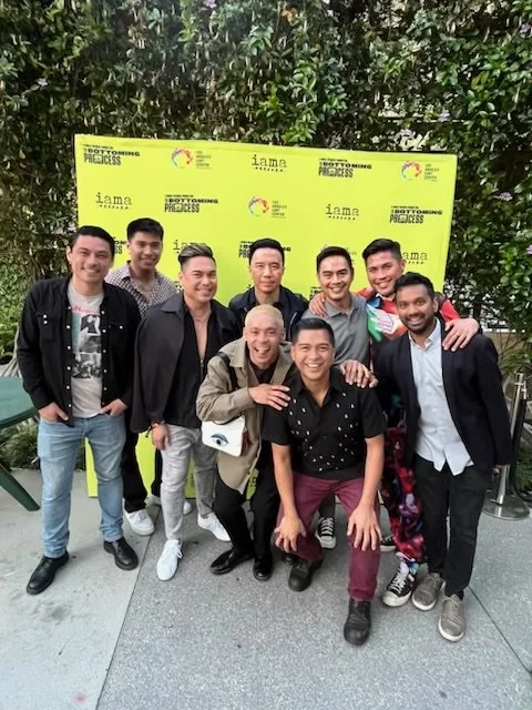 Group of nine young men posing outdoors in front of a bright yellow background with logos and text related to the incoming process, trees visible behind the background, all smiling.