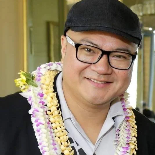 A man wearing glasses, a black cap, and a dark jacket, smiling, with a colorful lei around his neck.