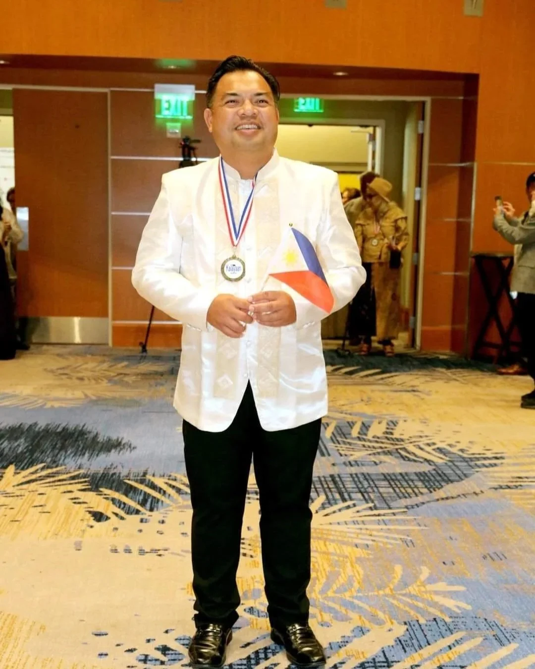 Man in a white traditional Filipino Barong shirt holding a Philippine flag and wearing a medal, standing in a conference hall.