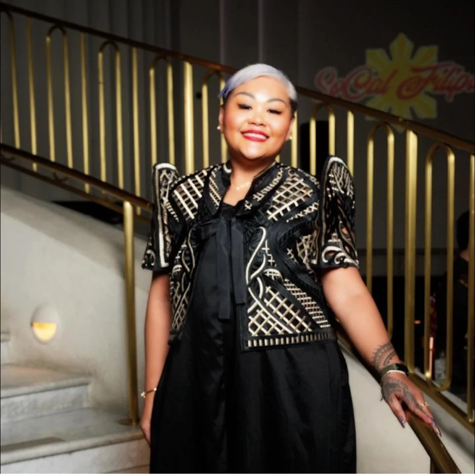 A smiling woman with short, silver hair wearing a black and gold patterned top standing on a staircase with a gold railing.