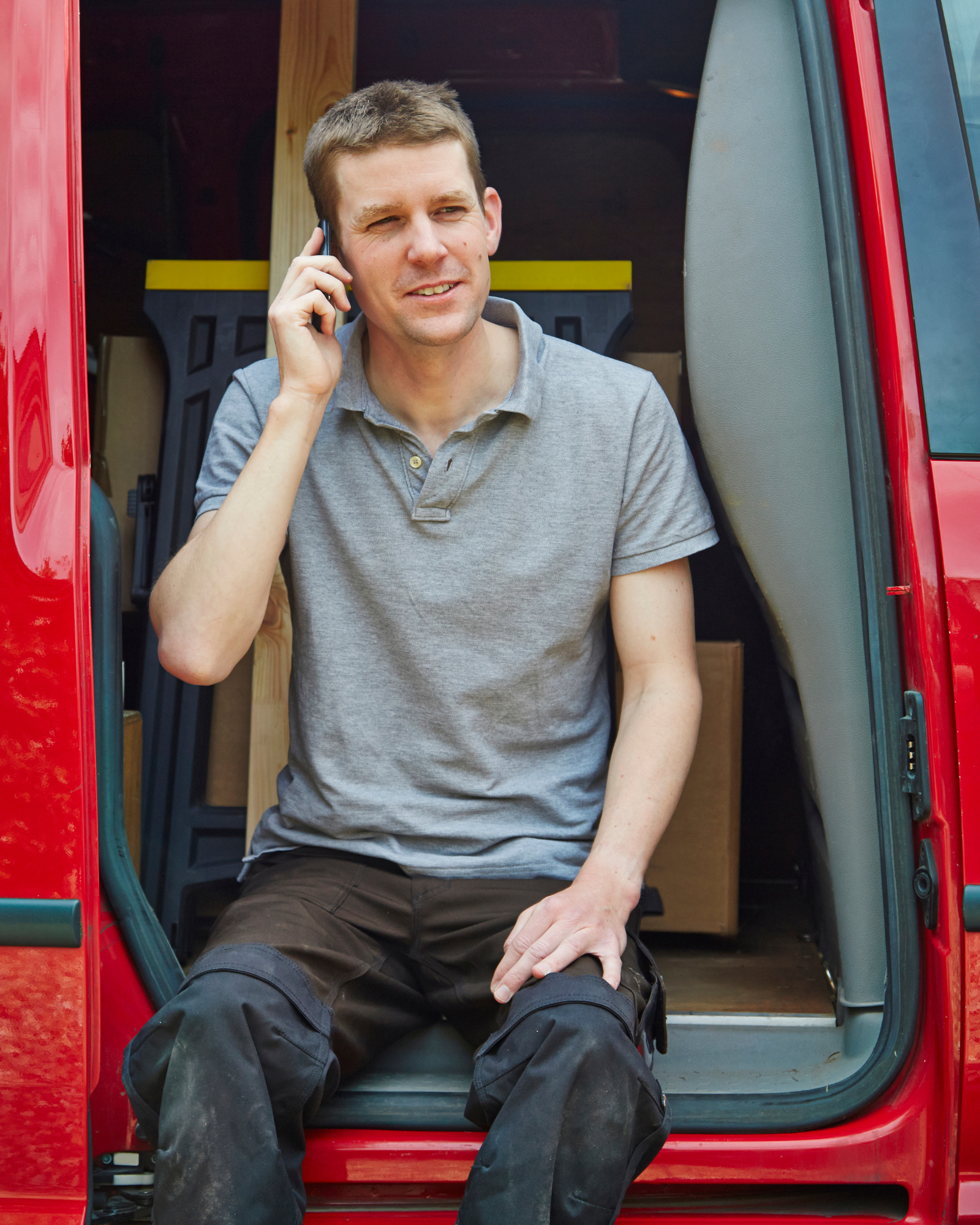 A young man sitting in the doorway of a red van, talking on a cellphone, wearing a gray polo shirt and black pants.