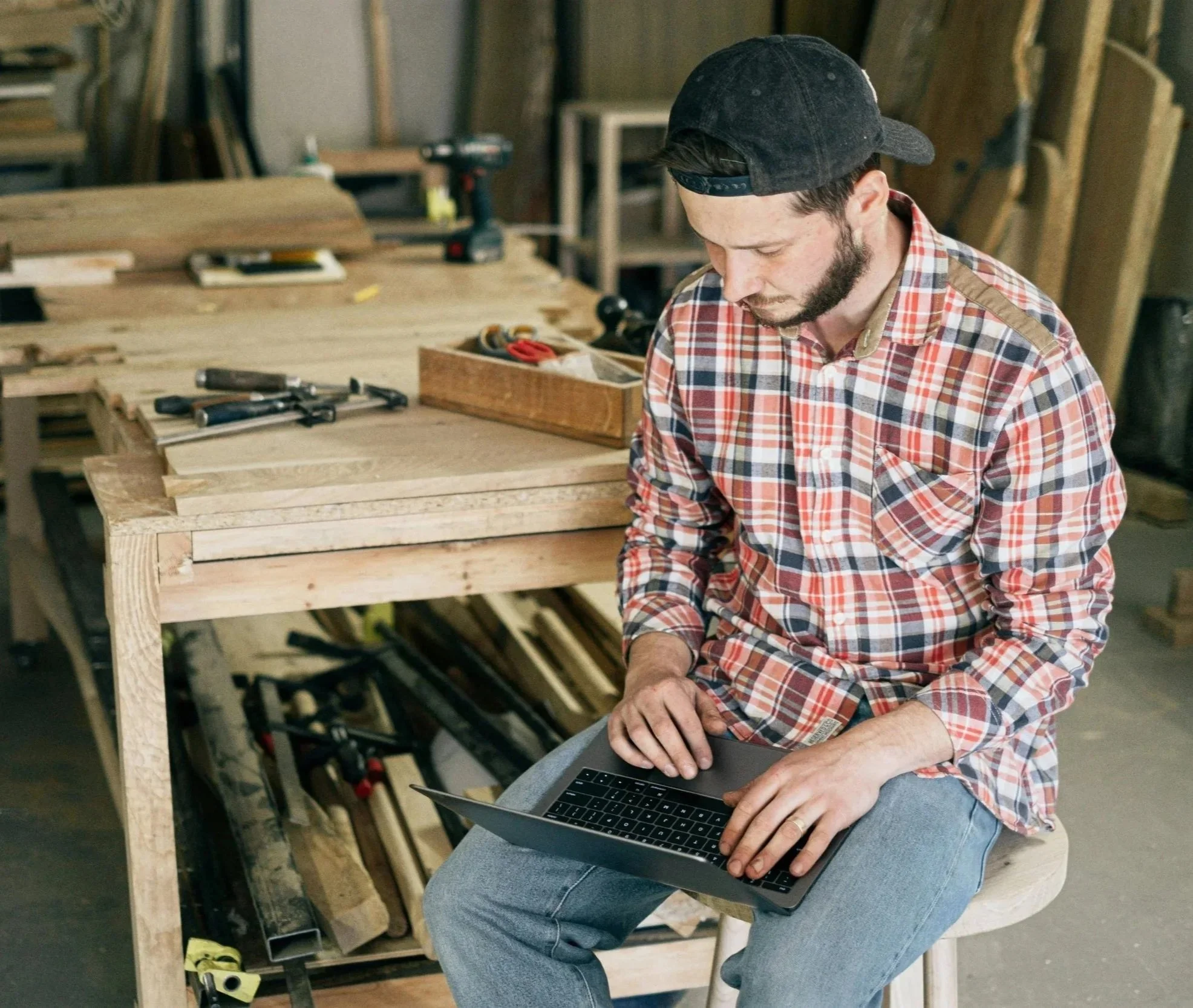 A man with a beard and wearing a black cap and a red plaid shirt sitting on a stool in a woodworking shop, working on a laptop placed on his lap, surrounded by woodworking tools and wood pieces.