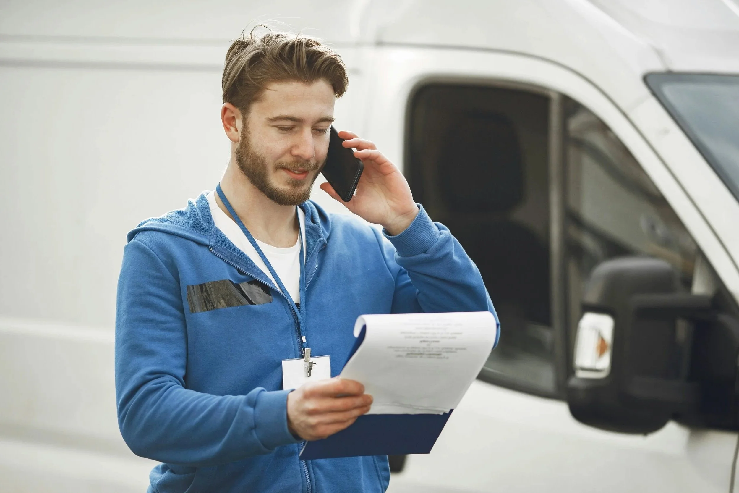 A young man with brown hair and a beard, wearing a blue hoodie, is talking on a cell phone while holding a clipboard and a pen, standing beside a white vehicle.