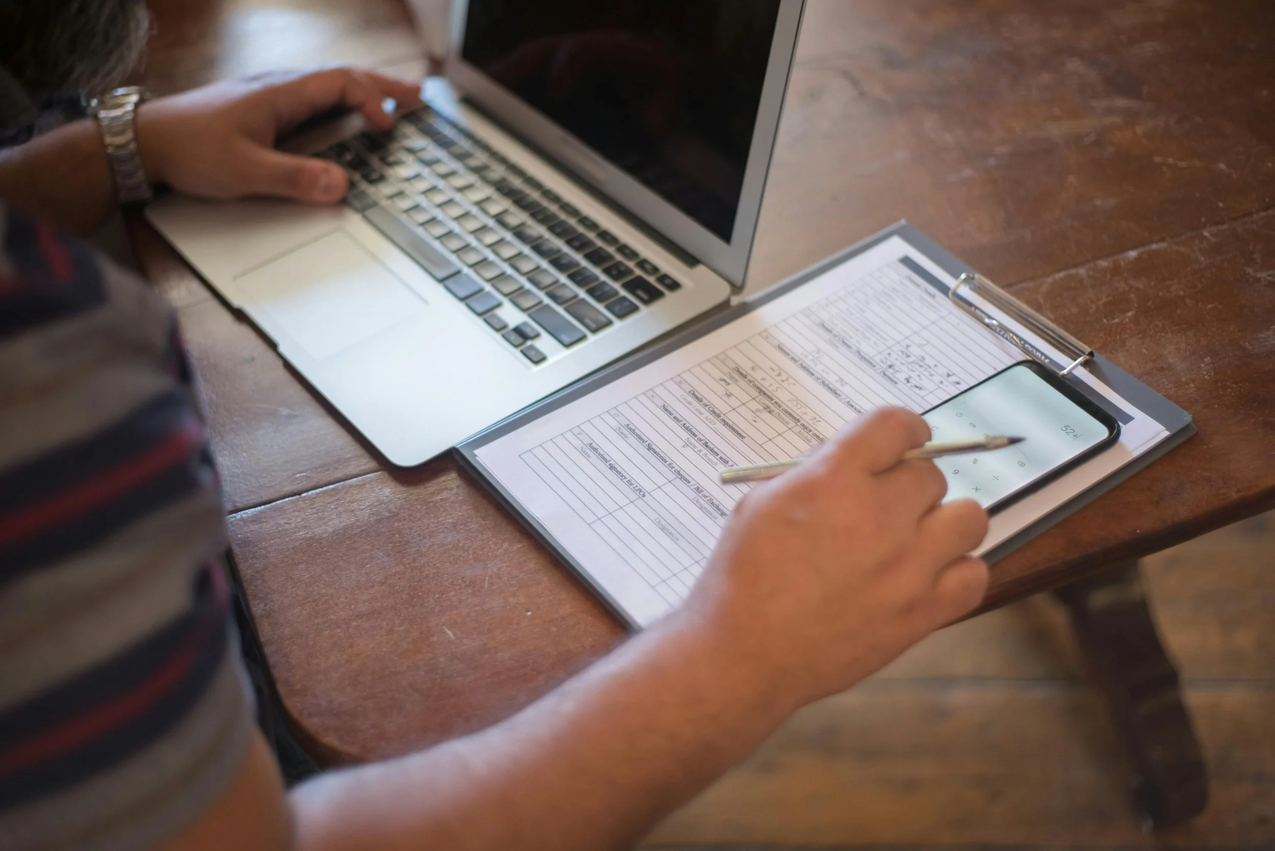 A person using a laptop and smartphone while working on a document on a wooden table.