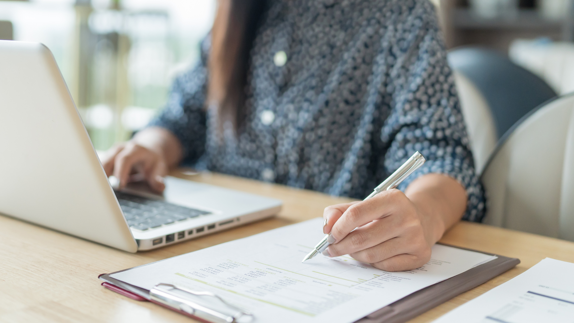 Person working at a desk with a laptop, writing on a paper, and holding a pen.