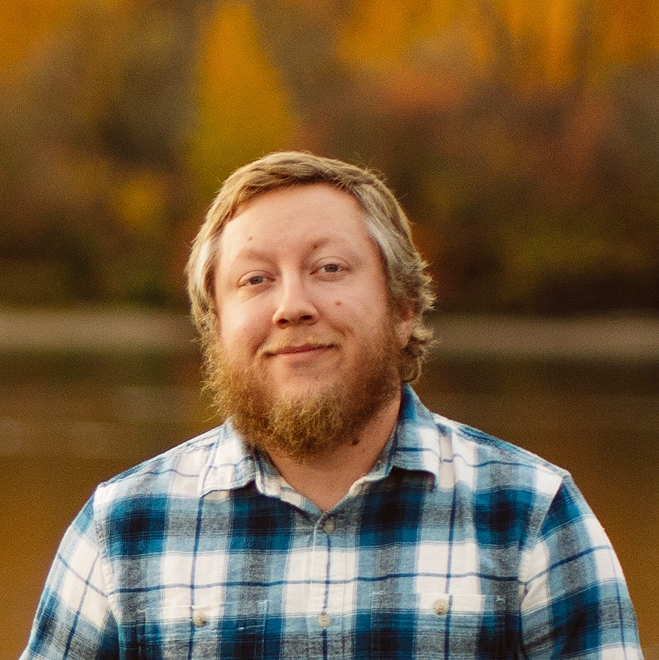 A young man with a beard and curly hair smiling outdoors, wearing a blue and white plaid shirt, with a background of autumn trees and a body of water.