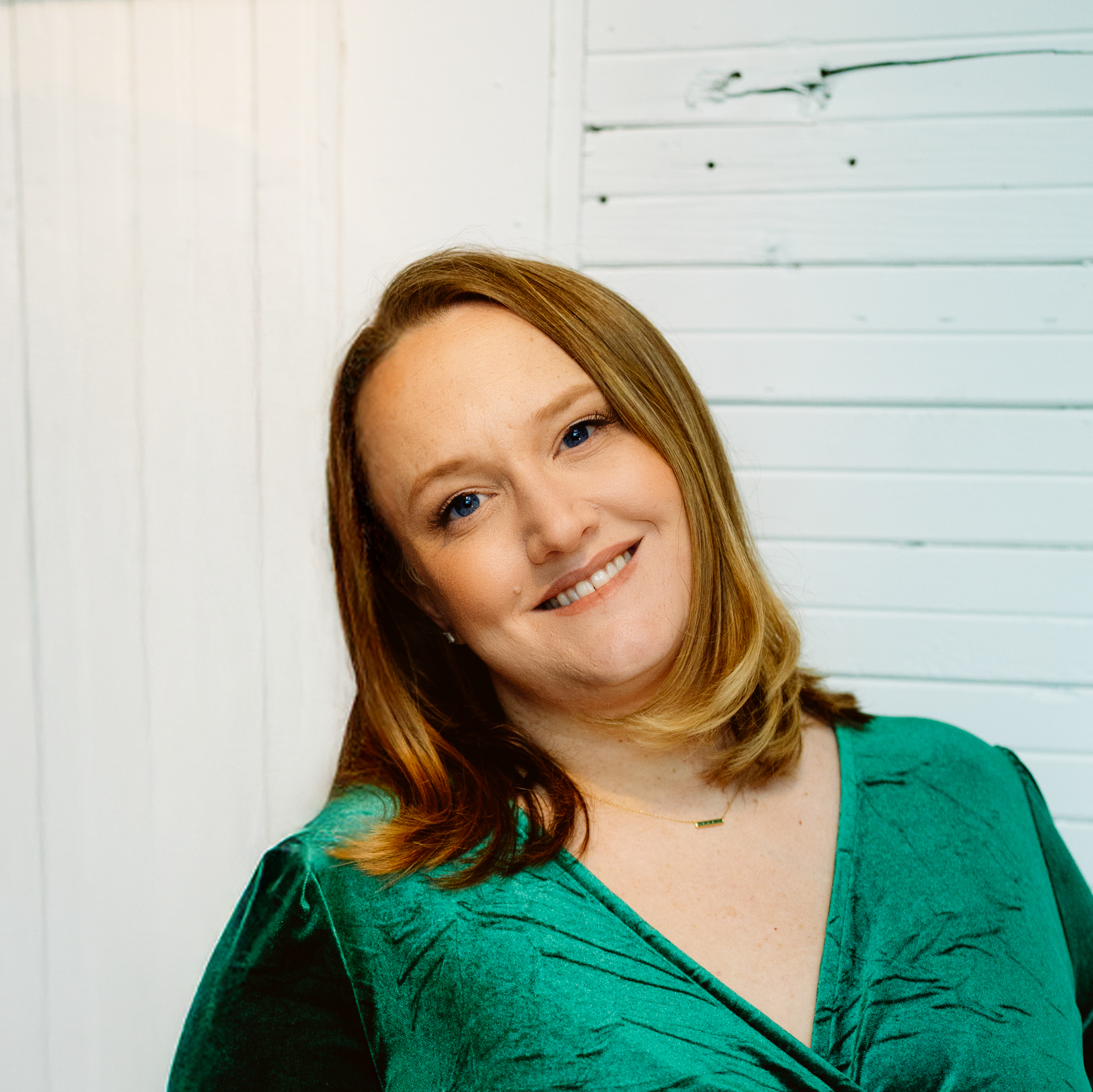 Close-up of a smiling woman with red hair, wearing a green top, standing against a white wooden wall.