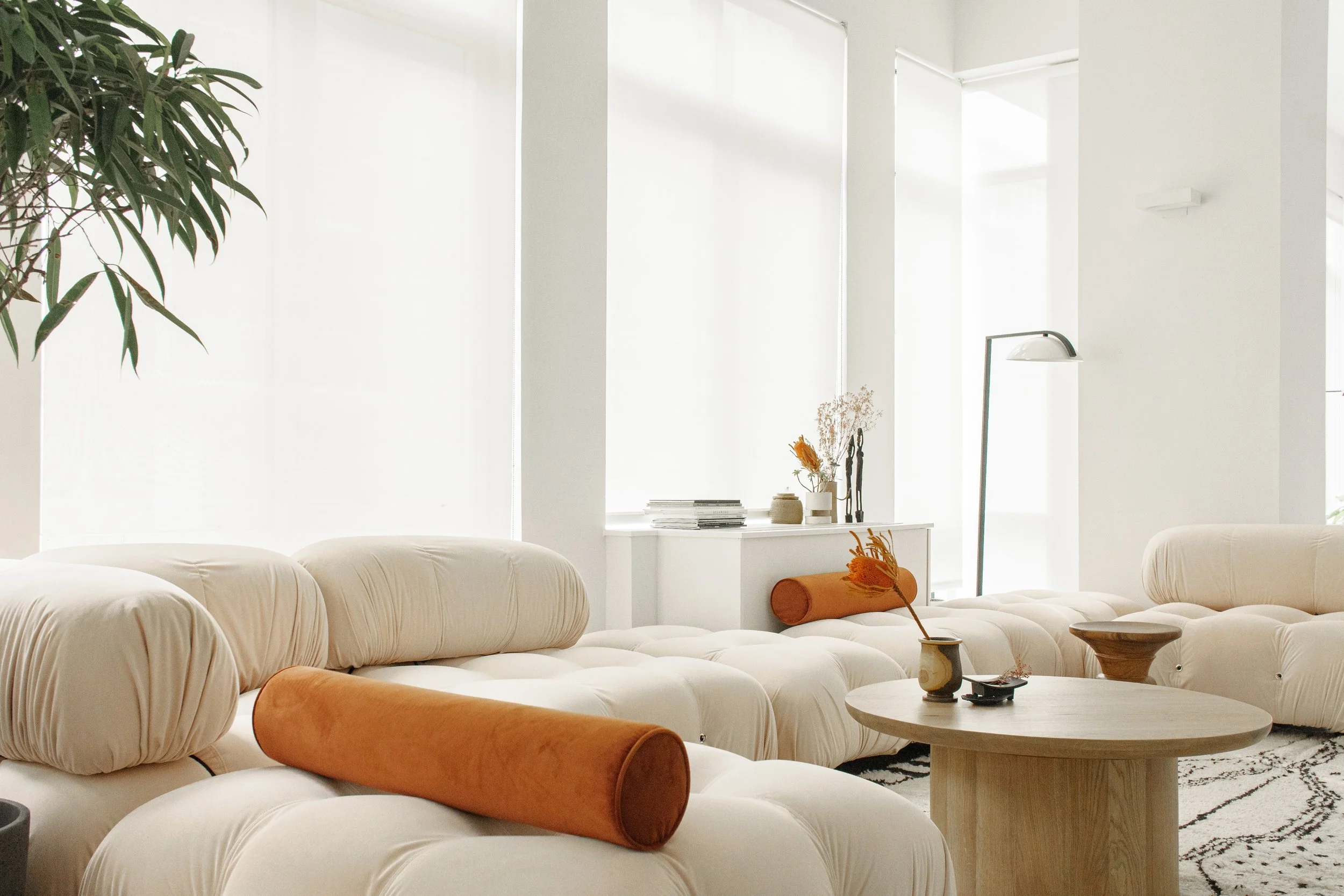 Minimalist living room with white tufted sofas, orange velvet bolster pillows, a round wooden coffee table, a modern floor lamp, and a white window sill with decorative vases and books, bright natural light.