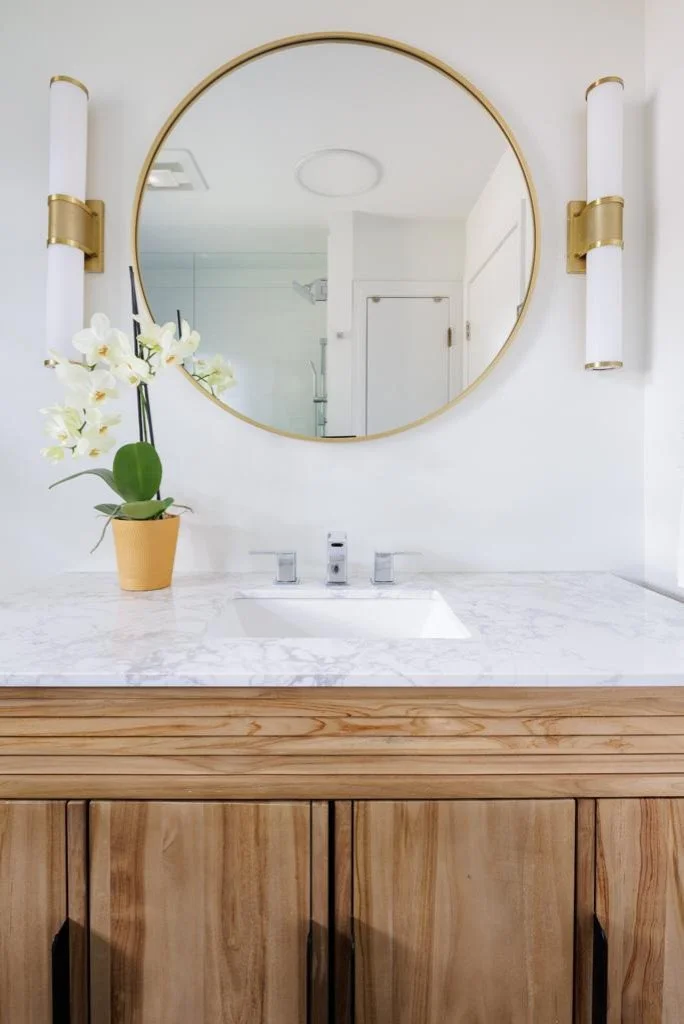 Bathroom vanity with a marble countertop, a single undermount sink with modern faucet, a large round mirror, wall-mounted light fixtures with white shades and gold accents, a potted white orchid, and wooden cabinet doors.