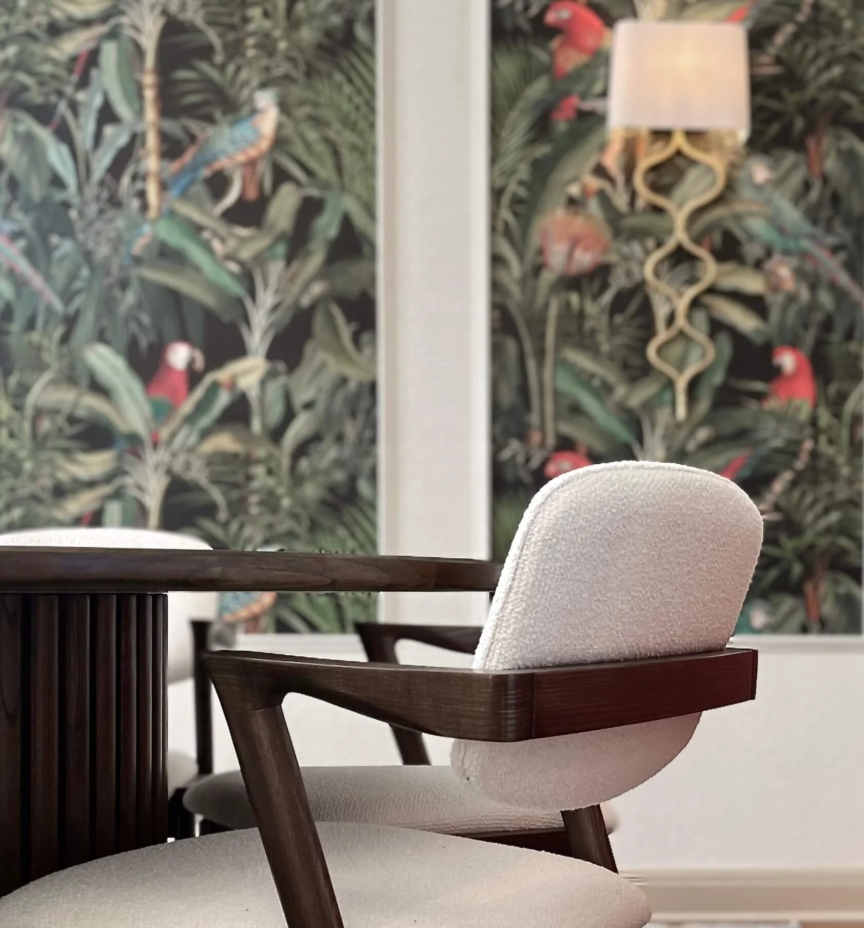 Interior of a room with a round wooden table and a modern white upholstered chair with wooden armrests, against a wall with tropical wallpaper featuring parrots and lush greenery, along with a sconce lamp.