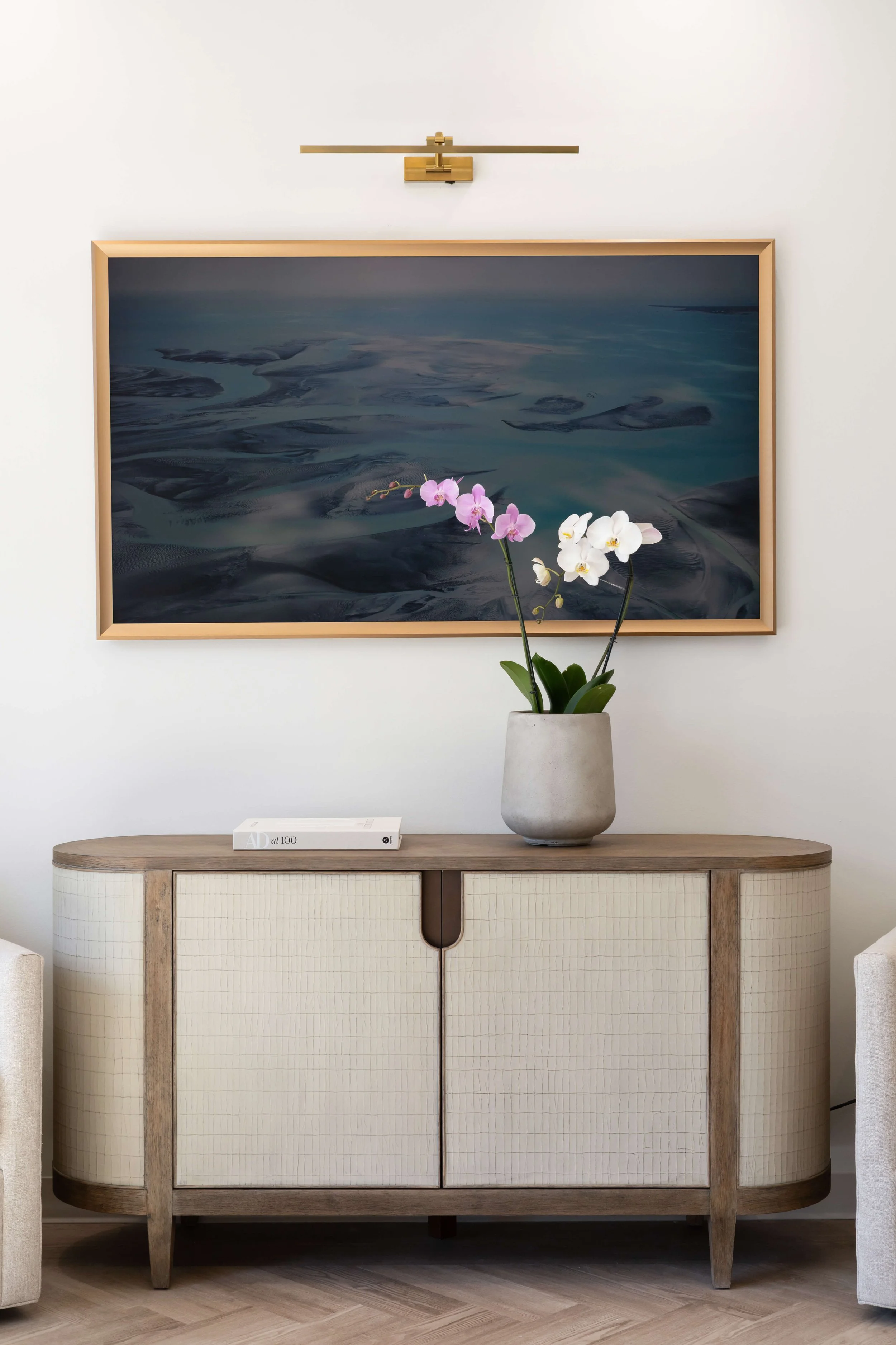 A wooden cabinet with two doors and a rounded top, floral artwork above, a vase with orchids on top, and a framed photograph or painting behind.