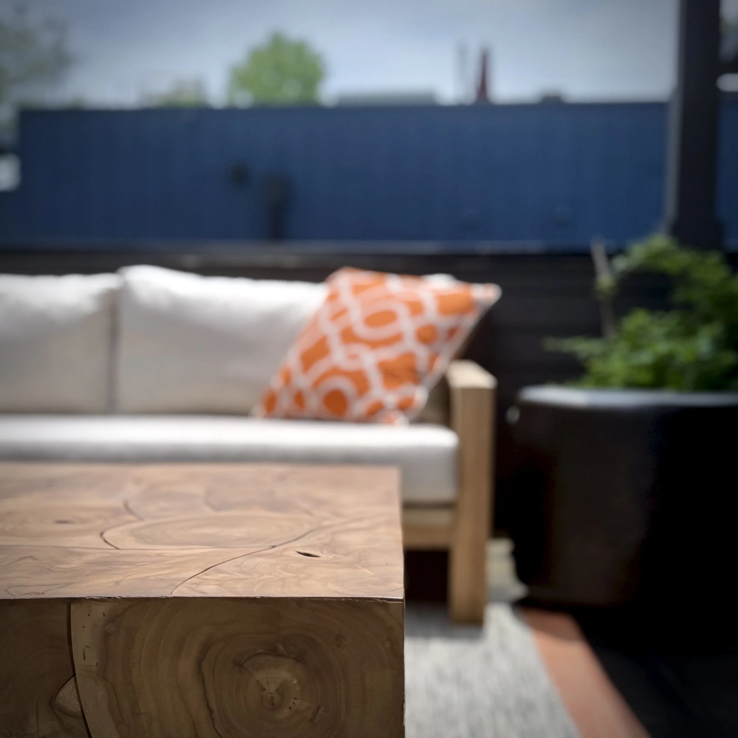 A close-up of a wooden coffee table with a natural wood grain, in front of a sofa with white cushions and an orange geometric pillow, on a patio with a dark wall and green plants, with a clear sky and a tree visible in the background.