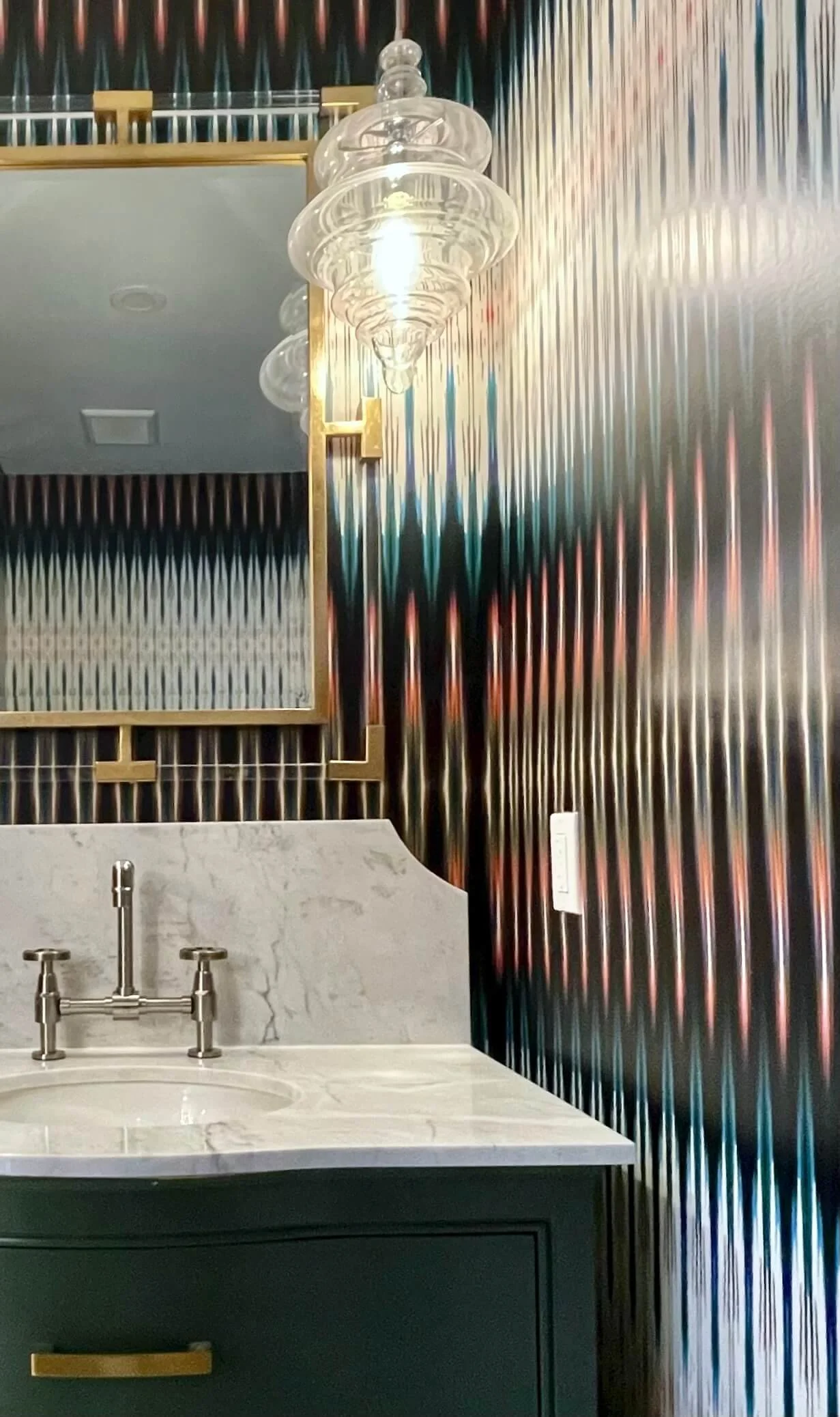 A bathroom vanity with a white marble countertop, a circular sink, and a silver faucet. Behind the vanity is a decorative wall with a metallic, rainbow-colored, ripple-texture finish. Above the vanity, there is a large rectangular mirror with a gold 