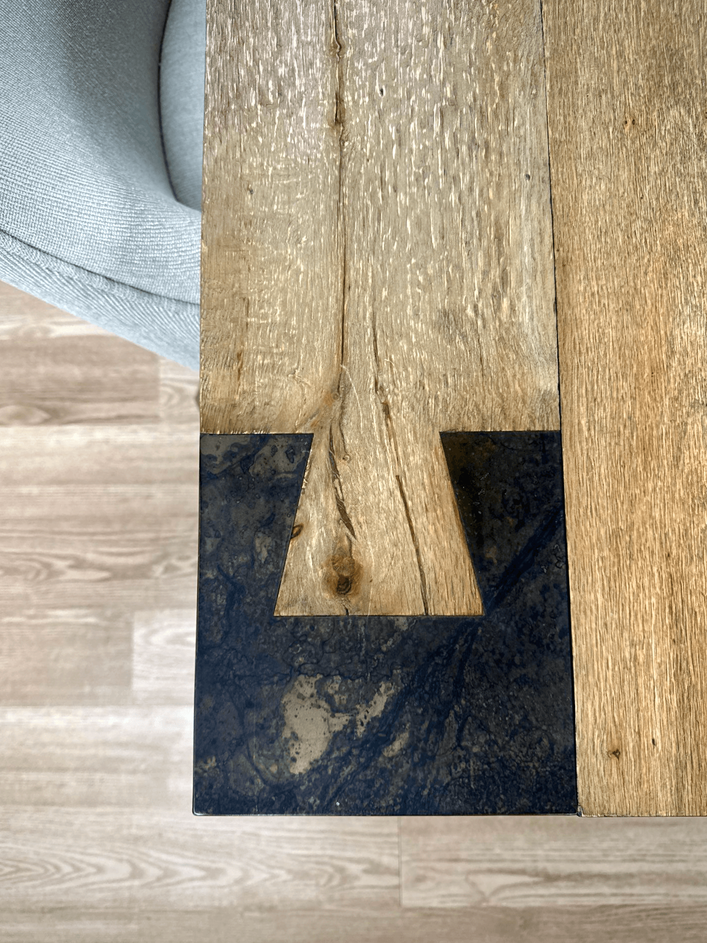 Close-up photo of a wooden table with black marble inlay and a corner of a gray upholstered chair visible.