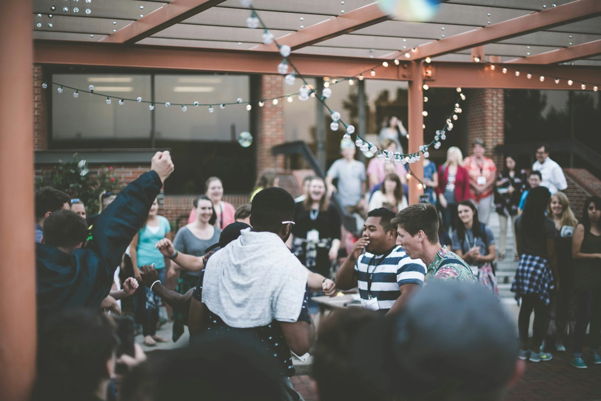 A diverse group of young people dancing and socializing outdoors under string lights, with some people standing in the background.