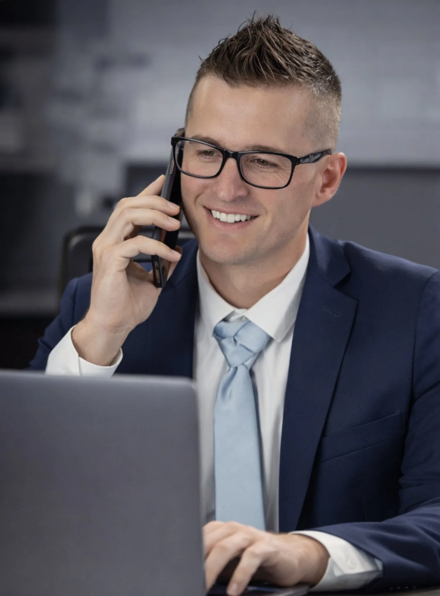 A man in a navy suit and glasses talking on a cell phone while working on a laptop.