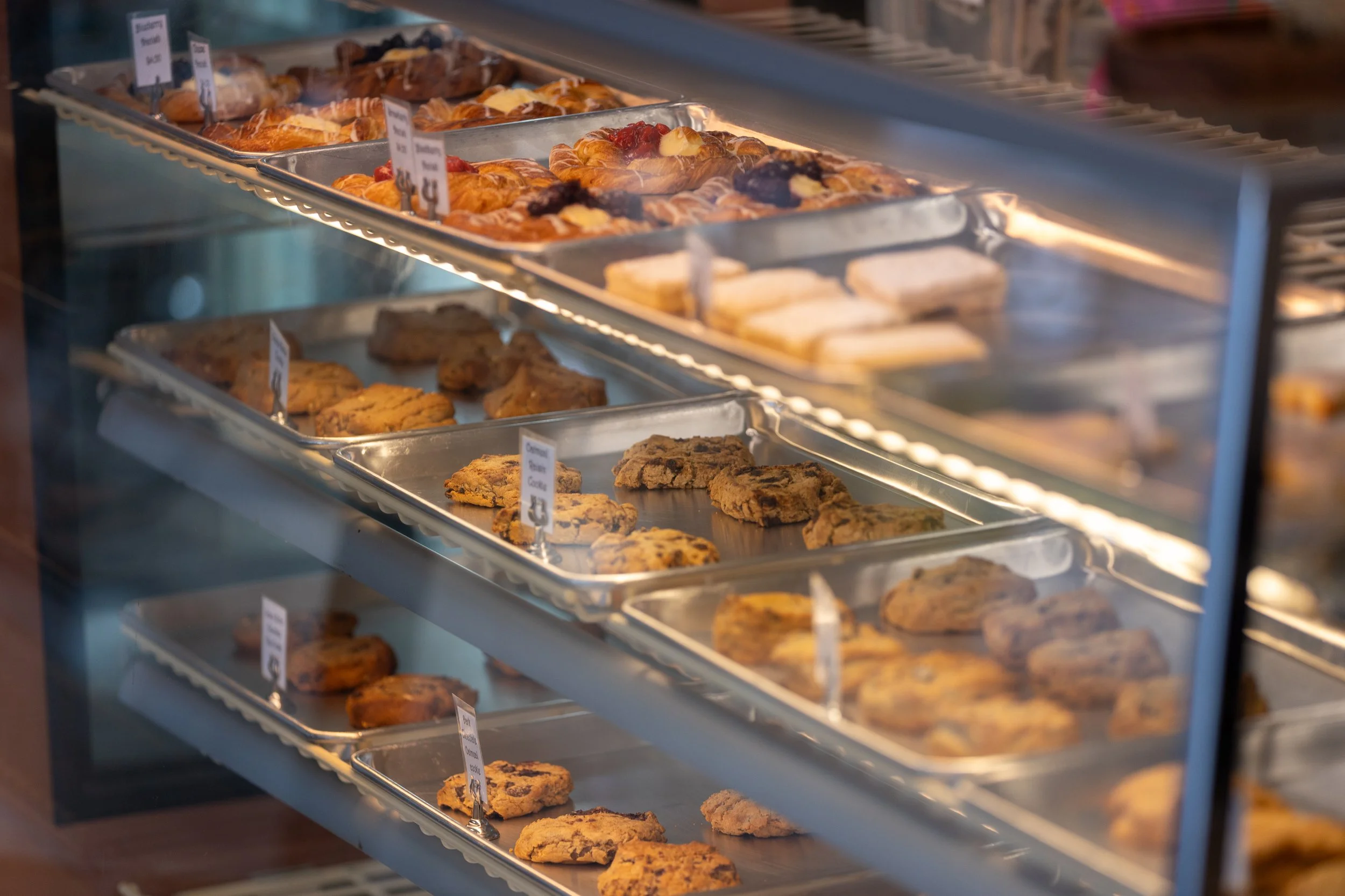 Display case filled with various types of baked cookies and pastries, each labeled with small signs, in a bakery or cafe shop.