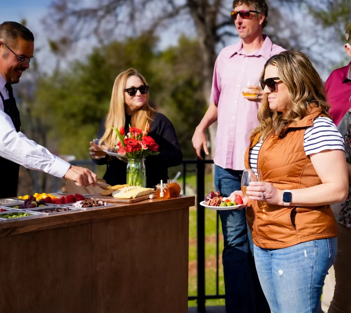 People at an outdoor gathering with a food table serving sliced fruits, cheese, and crackers, as one person serves food while others hold drinks and plates of food, with trees and clear skies in the background.