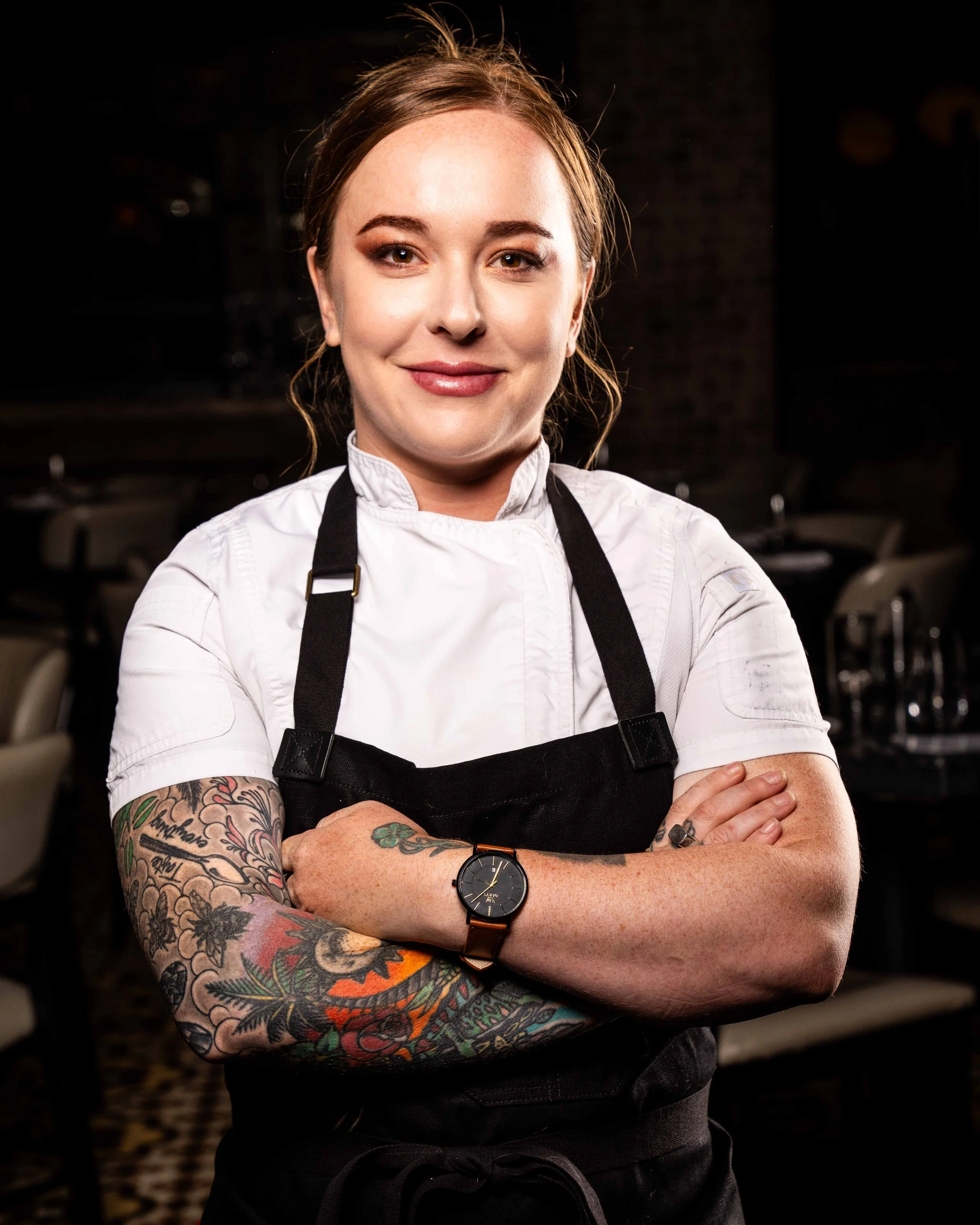 A female chef with tattoos on her arms, wearing a white chef's jacket and a black apron, standing confidently with arms crossed in a restaurant kitchen.