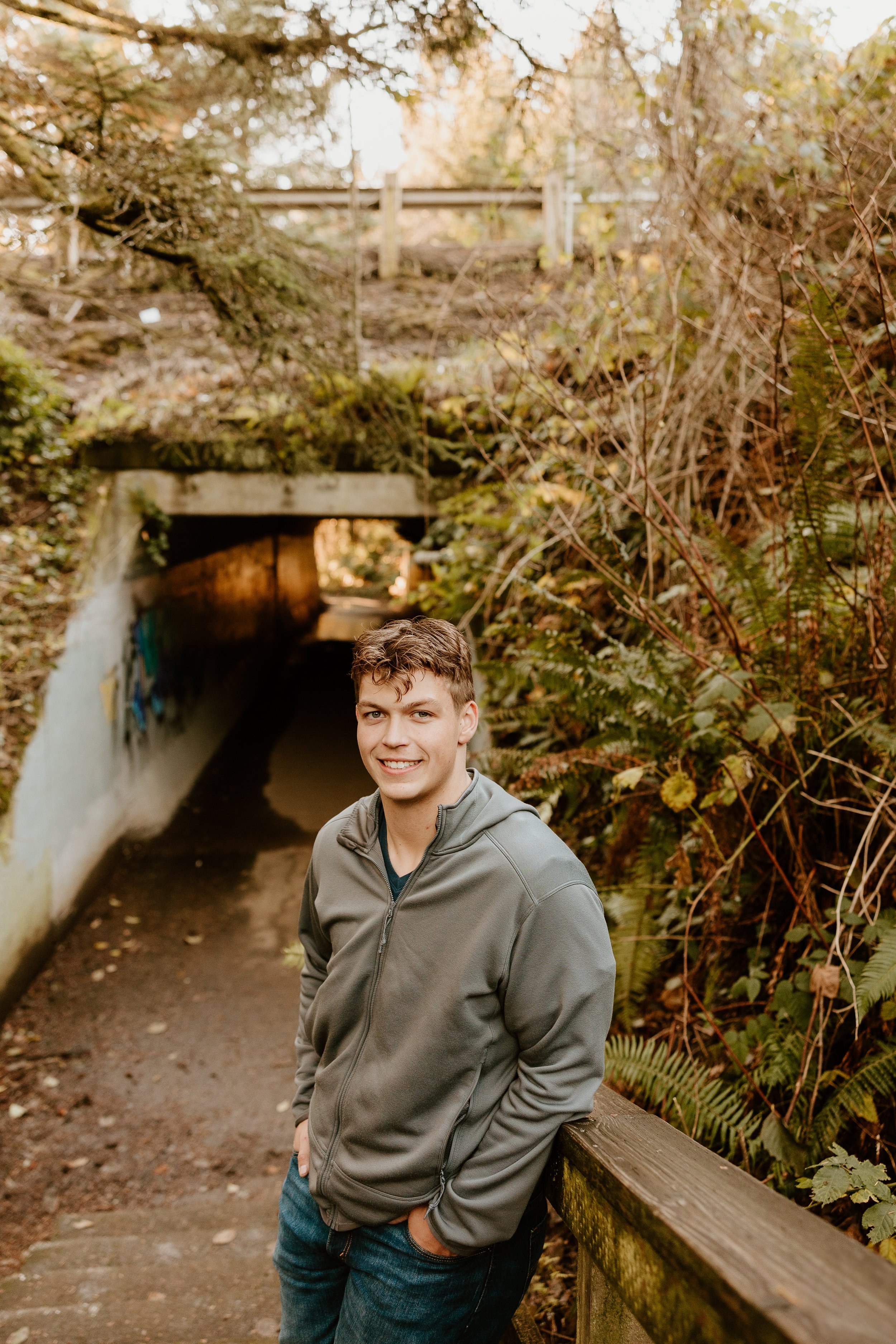 A young man in a gray jacket and blue jeans stands outdoors near a trail with a wooded backdrop, smiling at the camera.