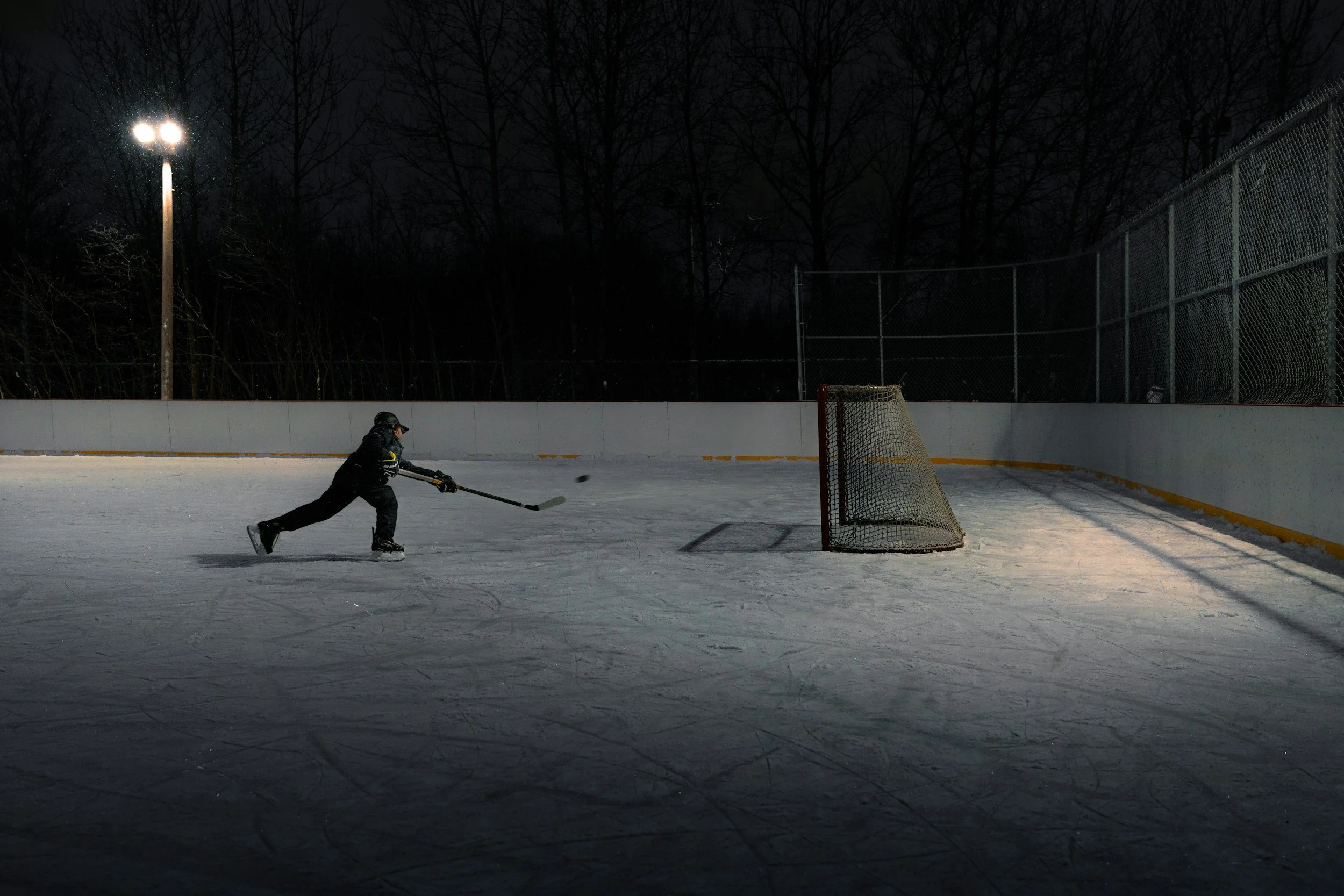 A young hockey player in black gear skating on an outdoor ice rink at night, with a hockey goal net nearby, surrounded by a chain-link fence and tall leafless trees, illuminated by a bright floodlight.