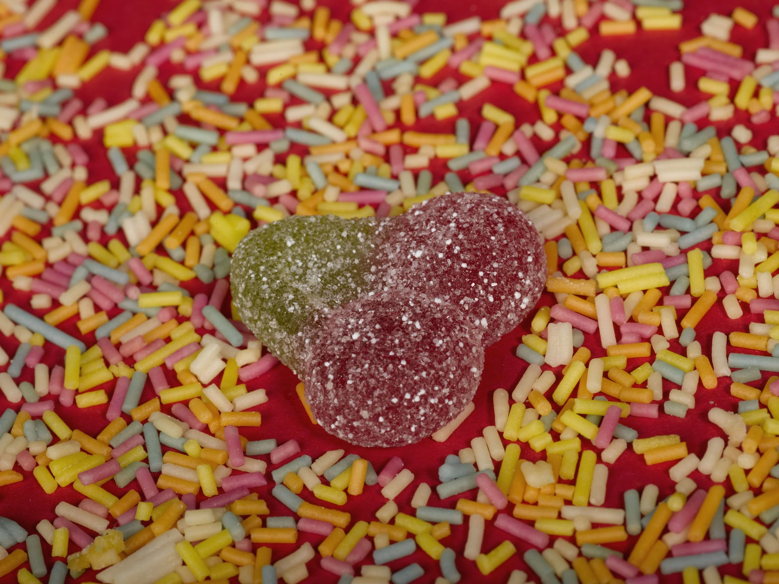 A heart-shaped gummy candy with sugar coating placed on a surface covered with multicolored sprinkles.