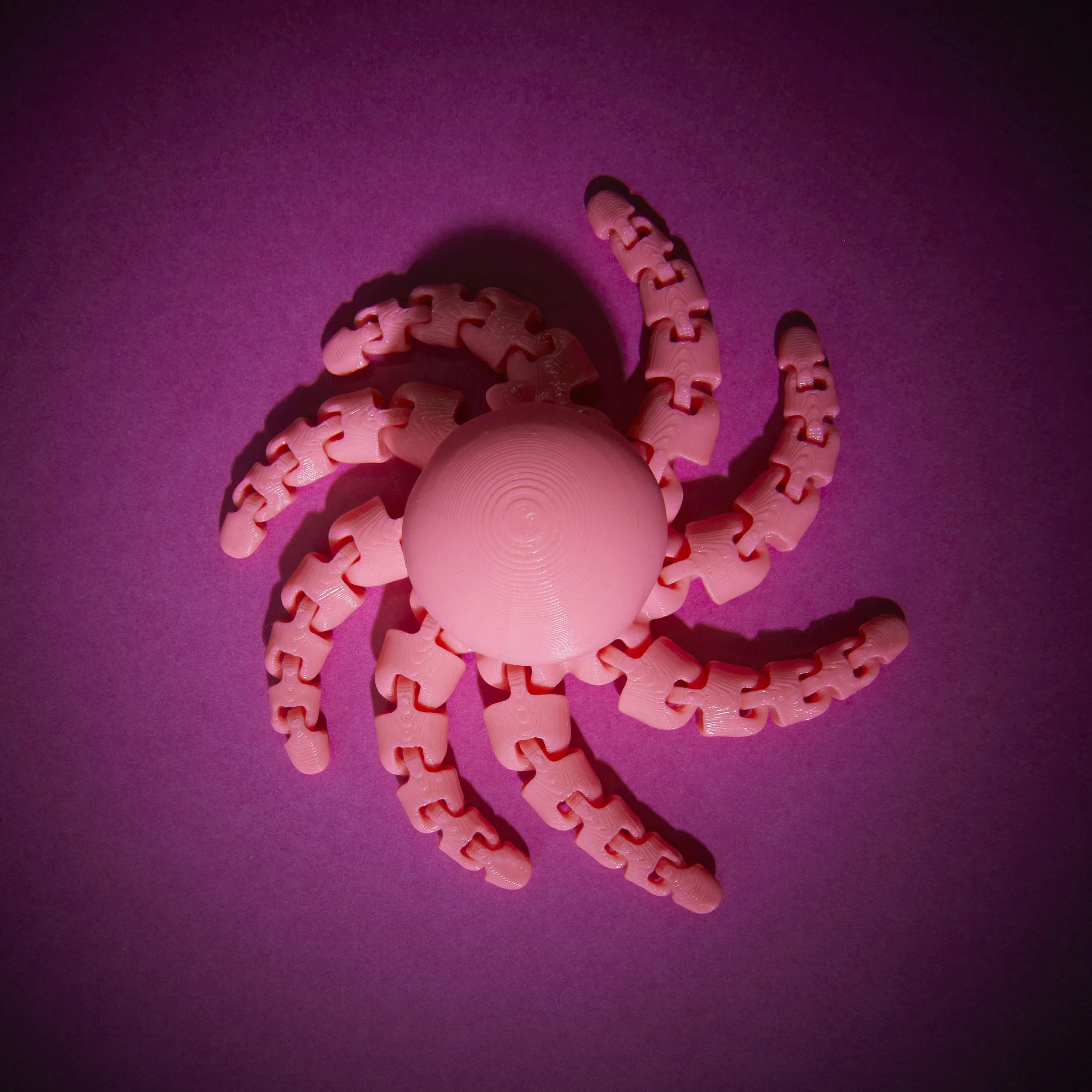 A pink plastic toy in the shape of a crab with a round body and six curved legs, placed on a purple surface with pink lighting.