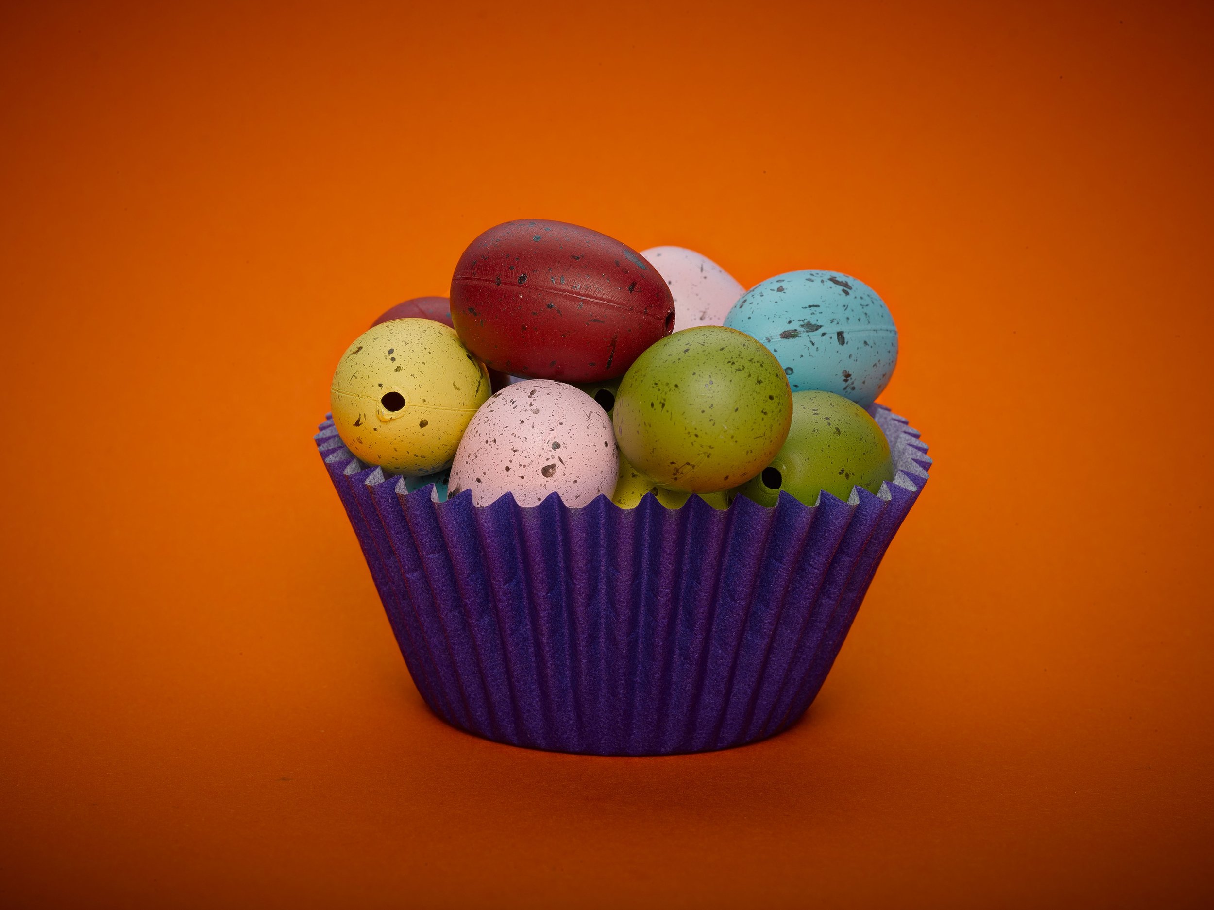 Colorful speckled Easter eggs in purple cupcake liner against orange background.