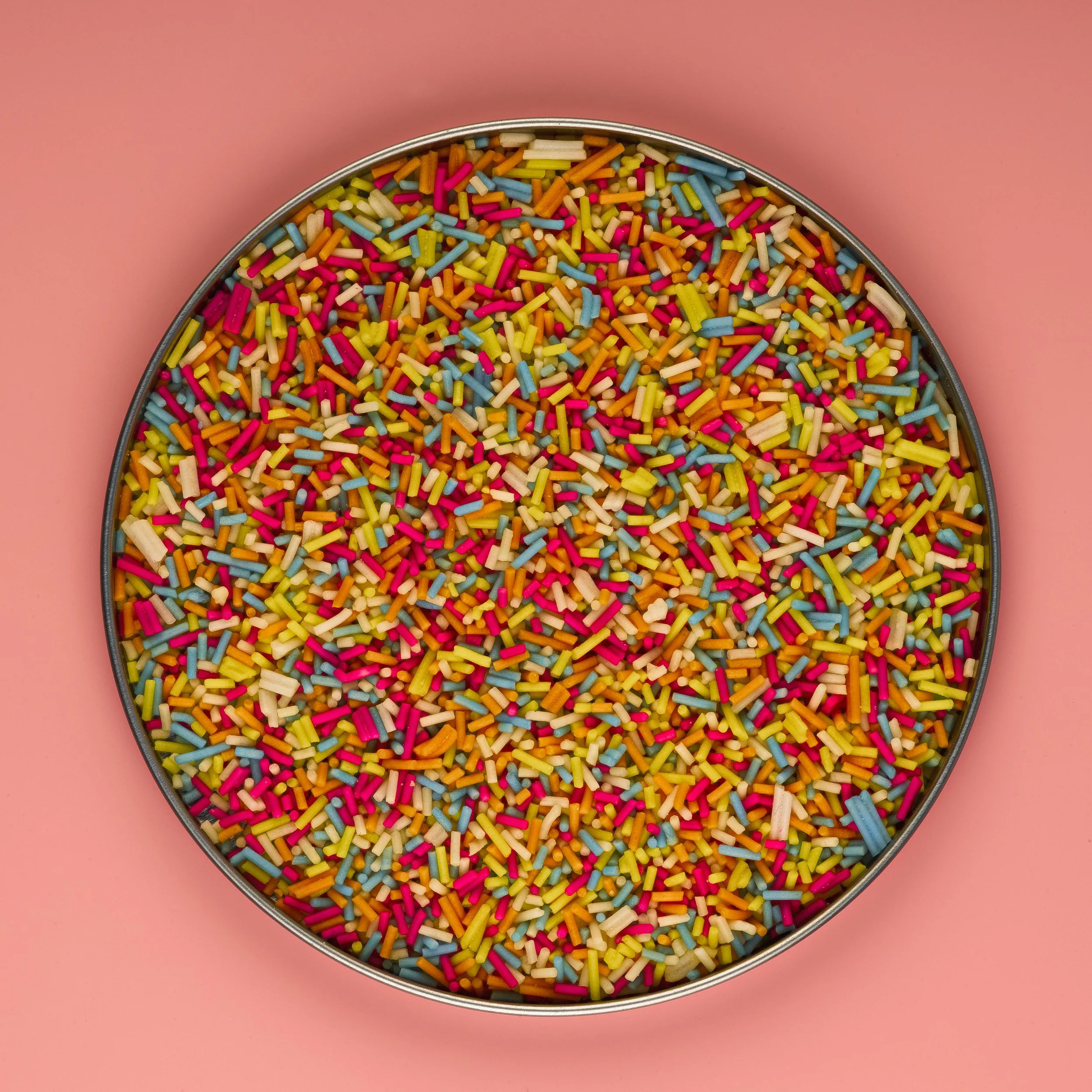 A round tin filled with multicolored rainbow sprinkles, placed on a pink background.