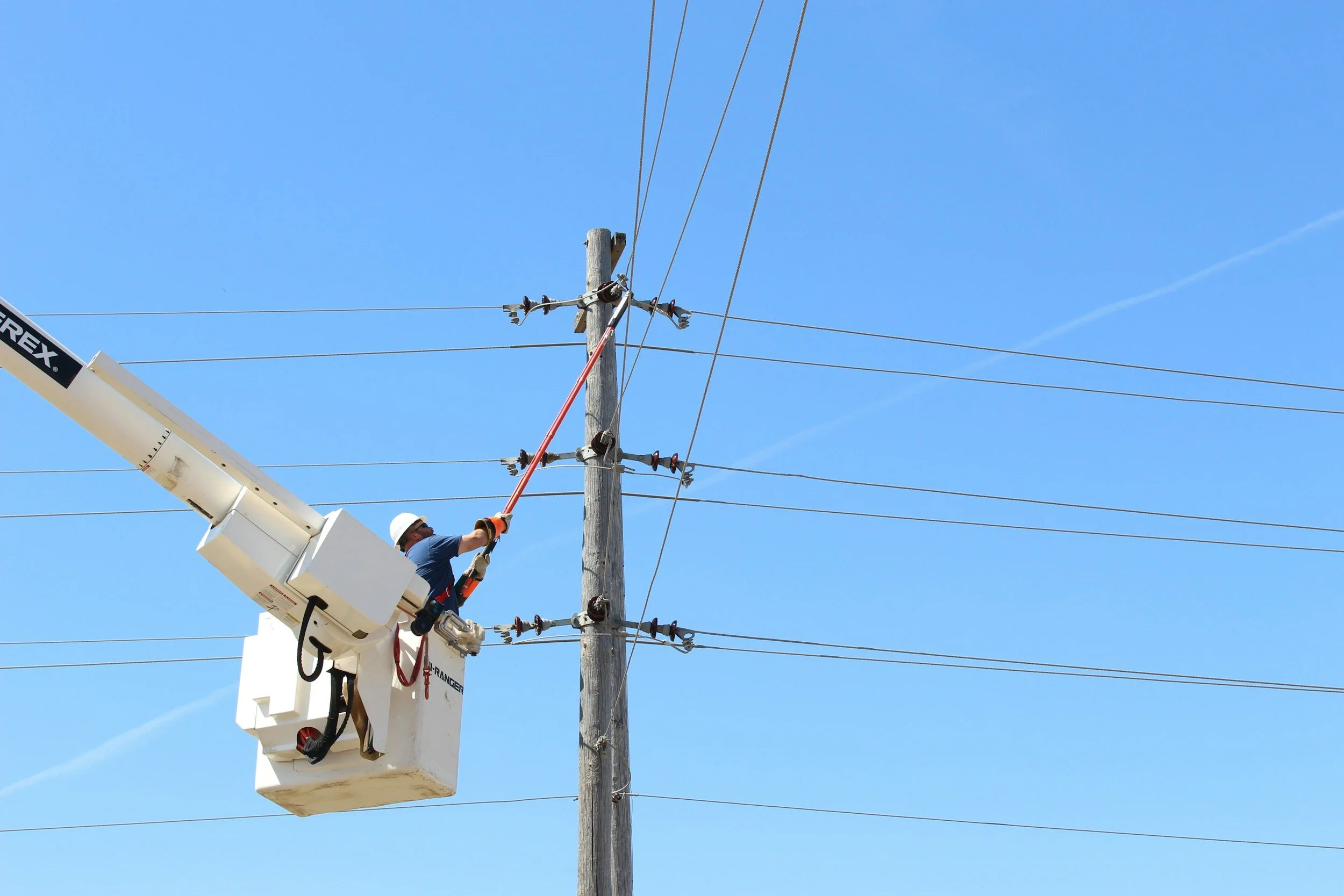A worker in a bucket lift working on power lines on a utility pole under a clear blue sky.