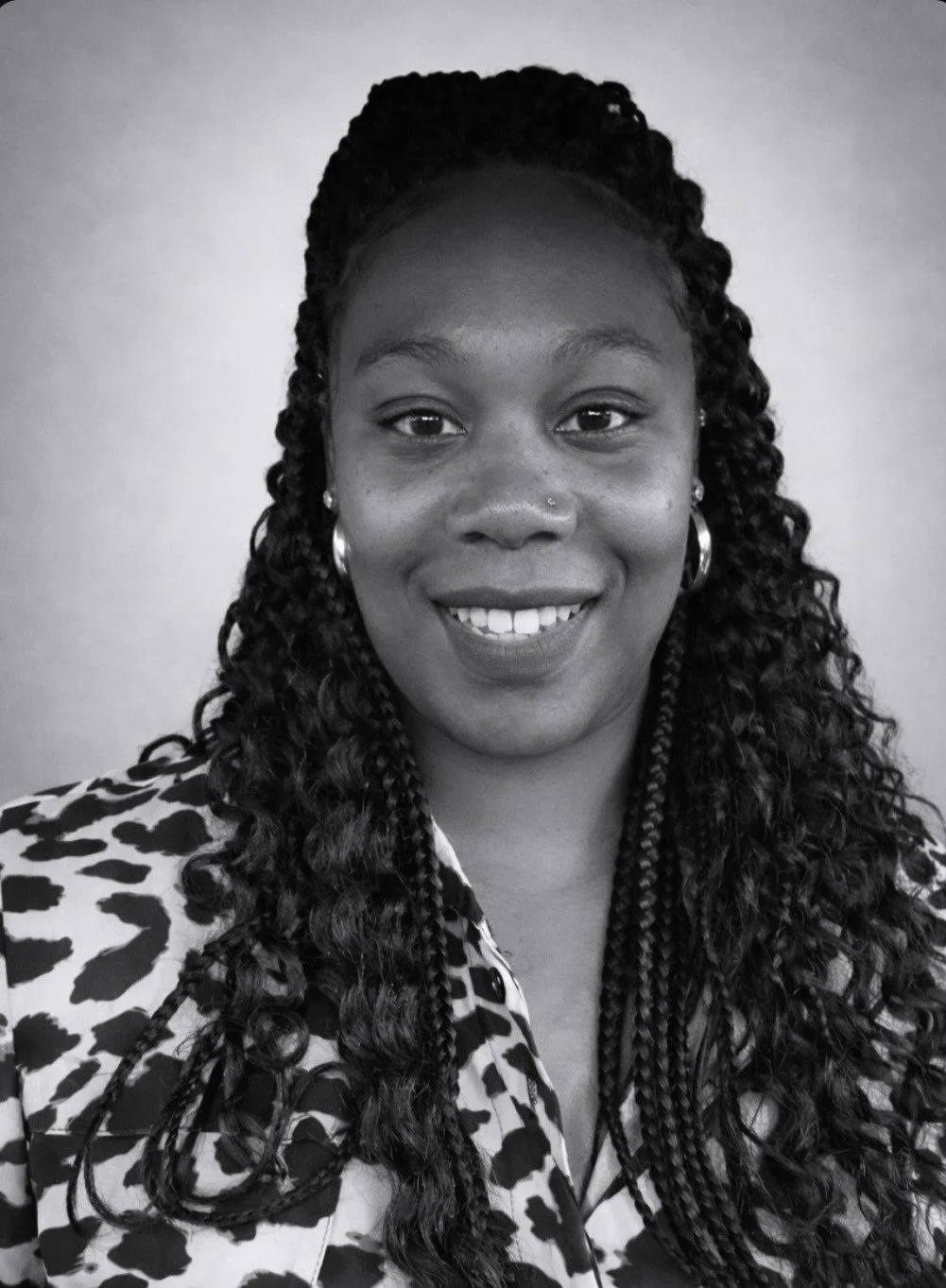 Black and white portrait of a woman smiling, with long, curly hair, wearing earrings, a small nose ring, and a patterned shirt.